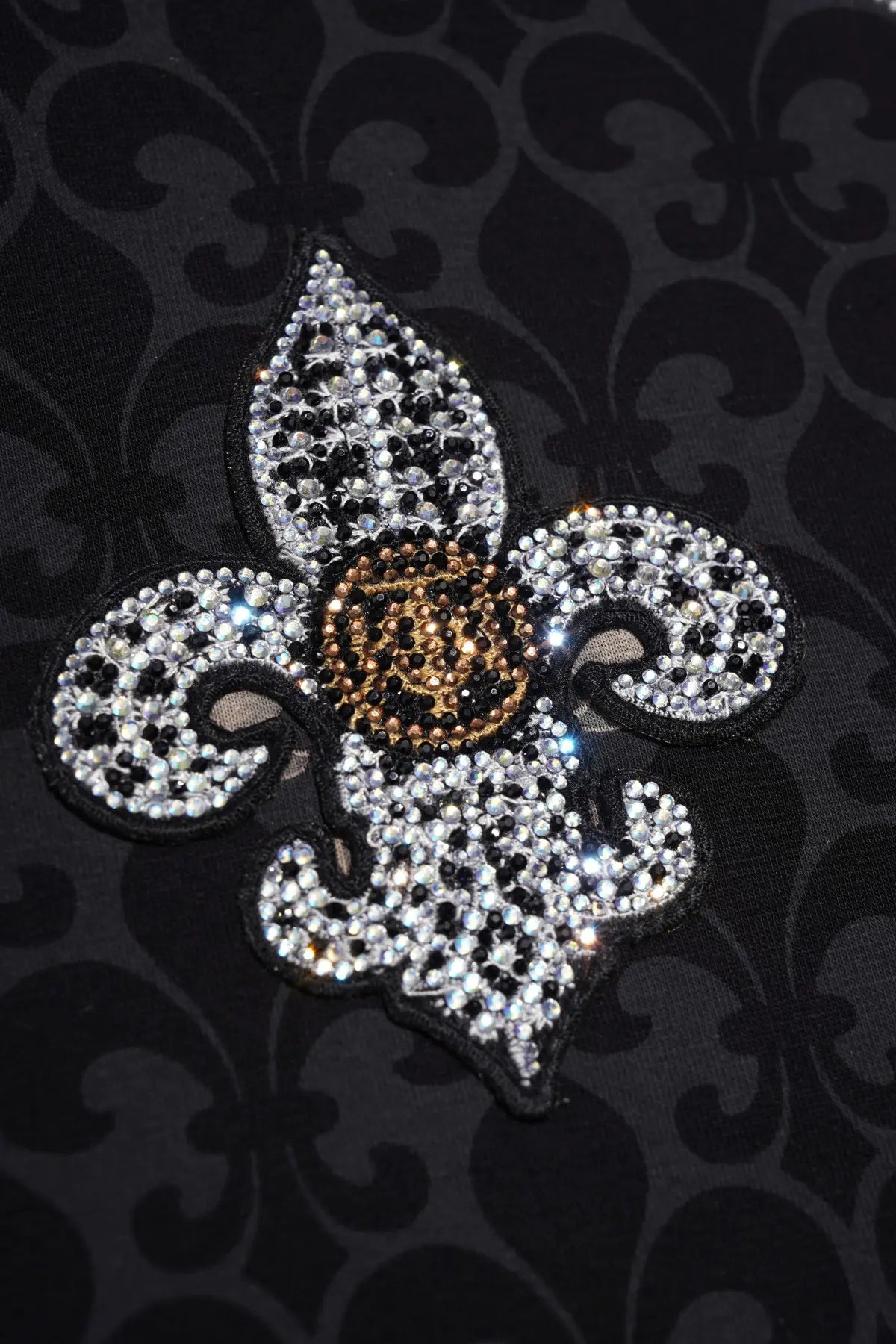 A fleur-de-lis style embellished patch or brooch with silver rhinestones, a black center, and a gold-toned textured middle circle, shown on a black damask fabric background. TCH Fleur-de-Lis Pattern Layered Long Sleeve T-Shirt