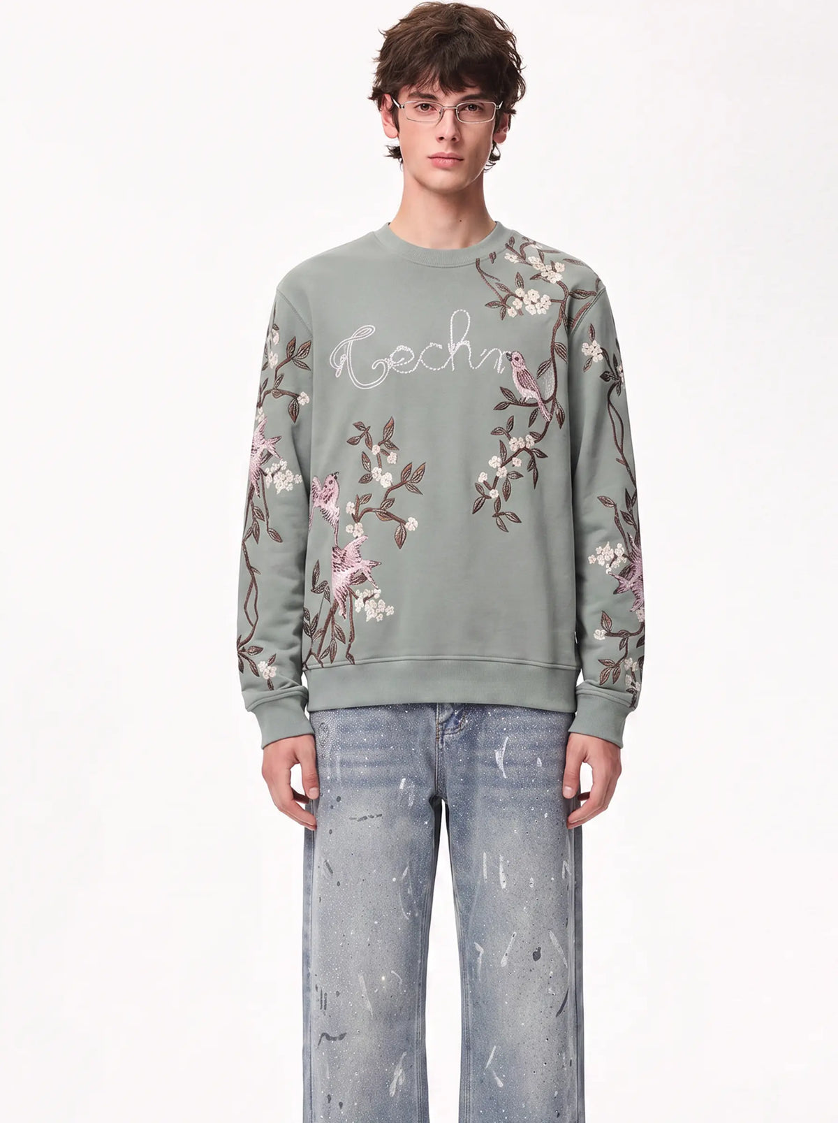 A model wearing a sage green crewneck sweatshirt with pink and white bird and floral embroidery, paired with light-wash jeans, against a plain white background. TCH Floral Bird Embroidery Oversized Sweatshirt