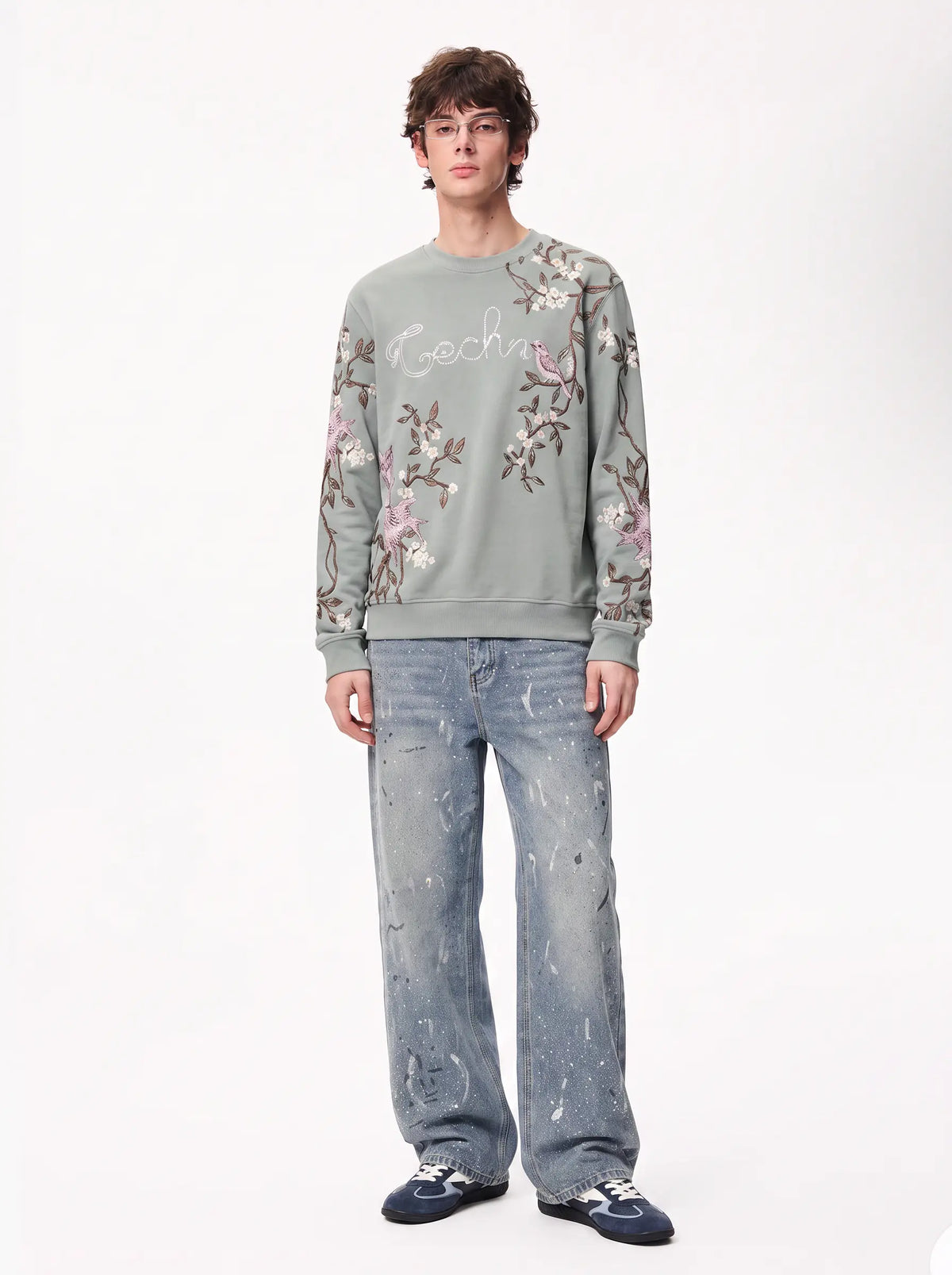 A male model wearing a sage green crewneck sweatshirt with pink and brown floral embroidery and white script lettering on the chest, paired with light-wash distressed jeans and dark blue sneakers, against a plain white studio background. TCH Floral Bird Embroidery Oversized Sweatshirt