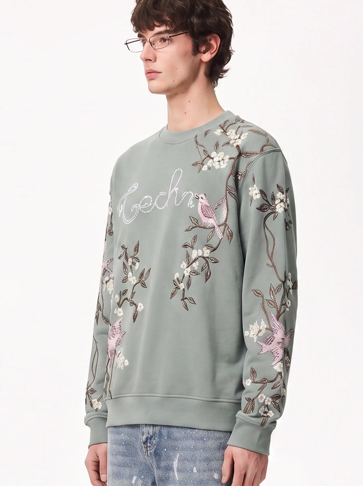 A man wearing a sage green crewneck sweatshirt with pink and white embroidered birds and floral motifs, paired with light blue jeans, against a plain white background. TCH Floral Bird Embroidery Oversized Sweatshirt
