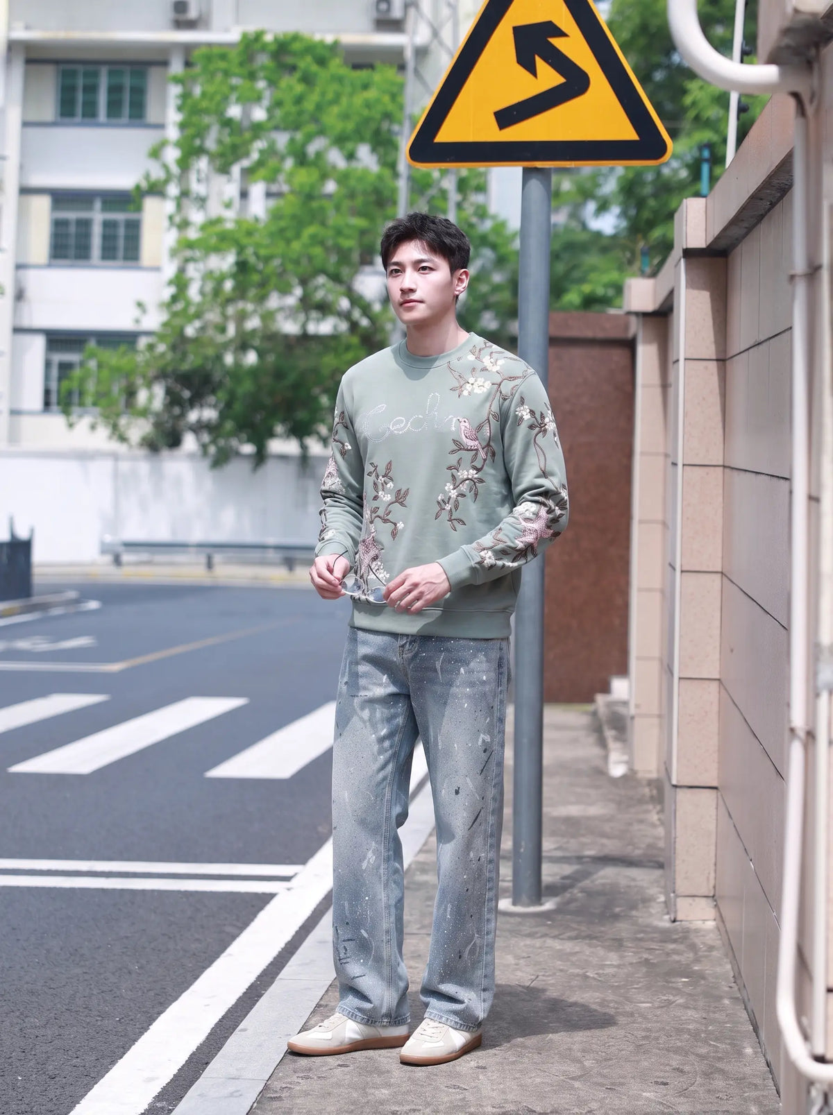 A man stands on a city sidewalk wearing a sage green crewneck sweatshirt with a floral pattern and light blue jeans, photographed outdoors near a street sign and building. TCH Floral Bird Embroidery Oversized Sweatshirt