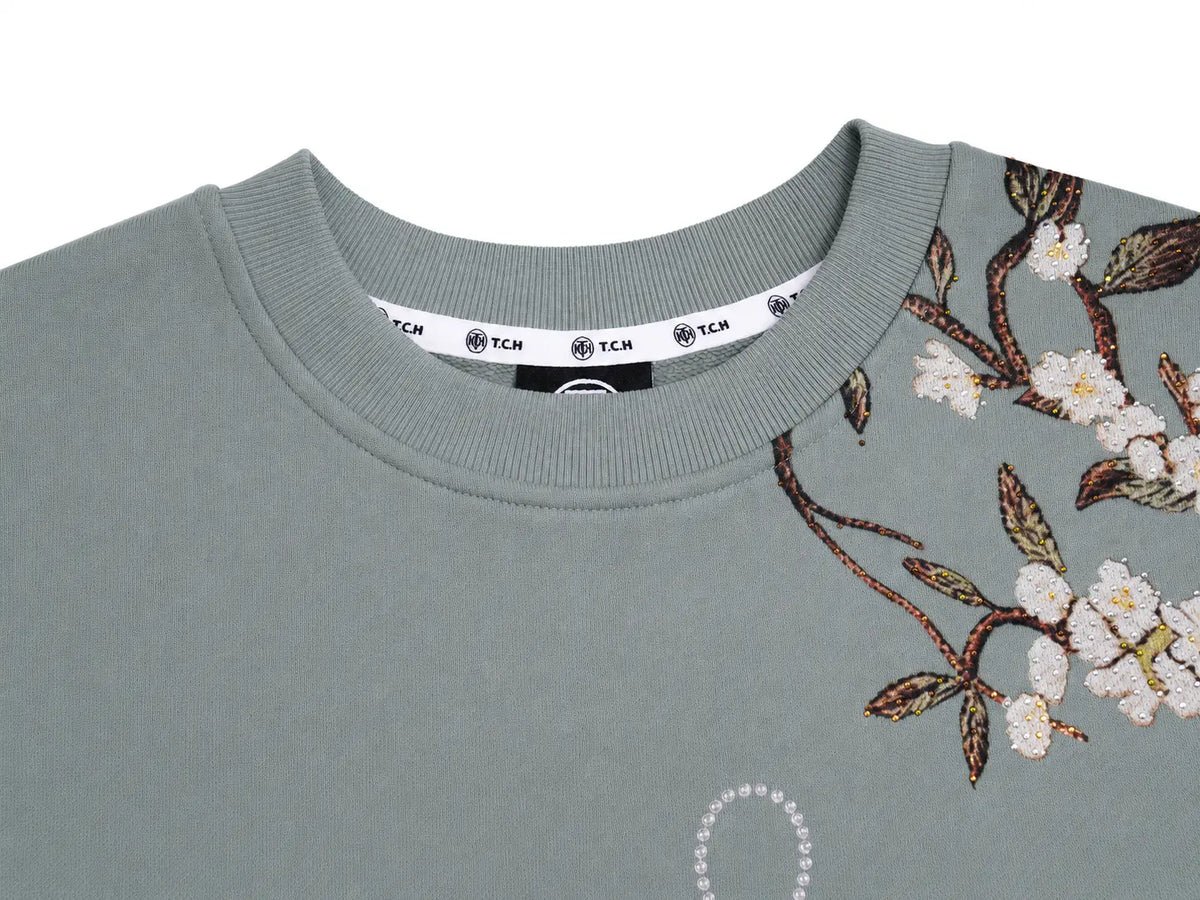 Close-up of a sage green crewneck sweatshirt featuring floral embroidery on the right chest with white blossoms and brown leaves, and a visible TCM neck label. TCH Floral Bird Embroidery Oversized Sweatshirt