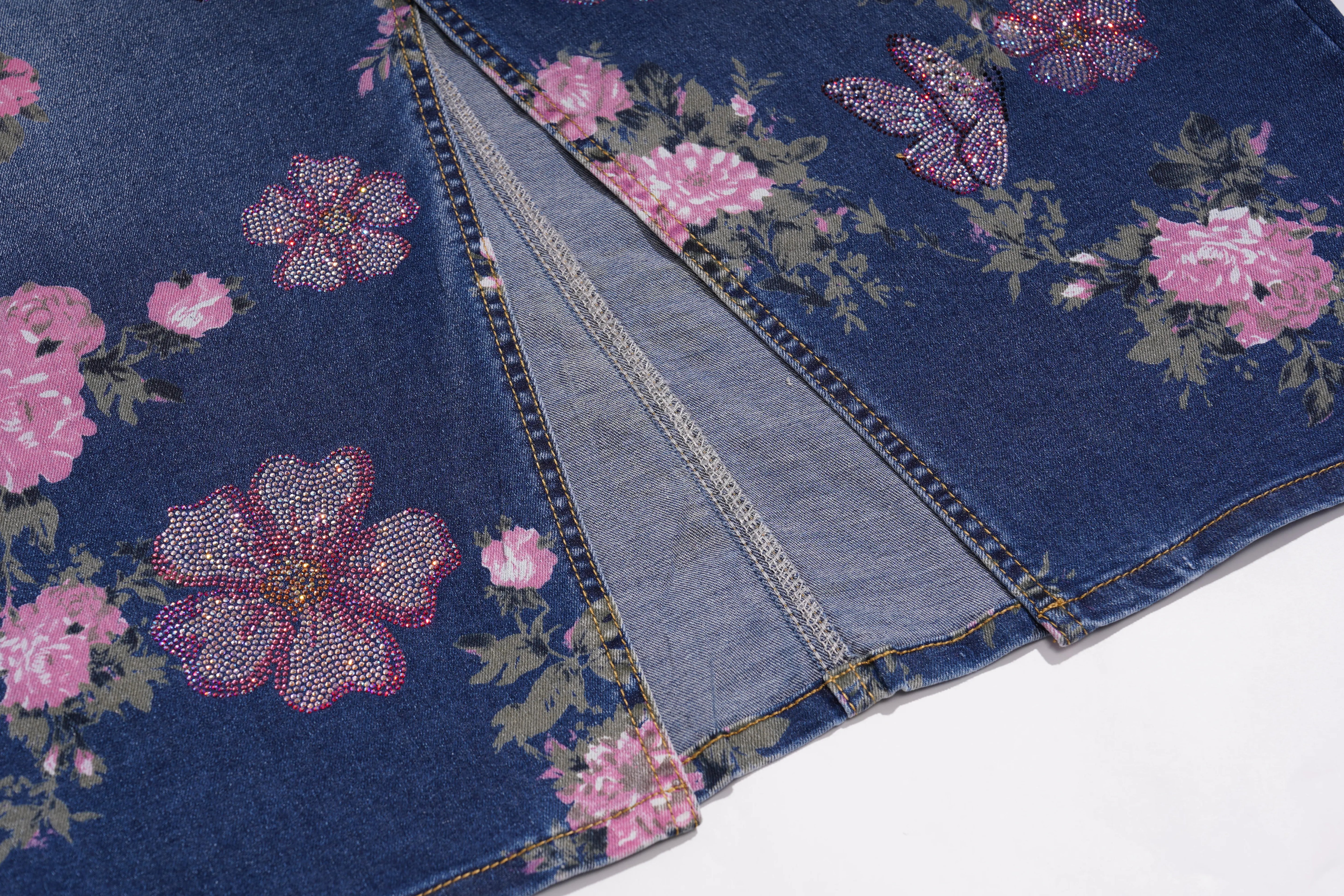 A close-up of a blue denim garment, likely a jacket or vest, featuring a pink and green floral print embellished with rhinestones. The image shows a diagonal split in the fabric, revealing the lighter blue inner lining. TCH Floral Butterfly Print Denim Midi Skirt