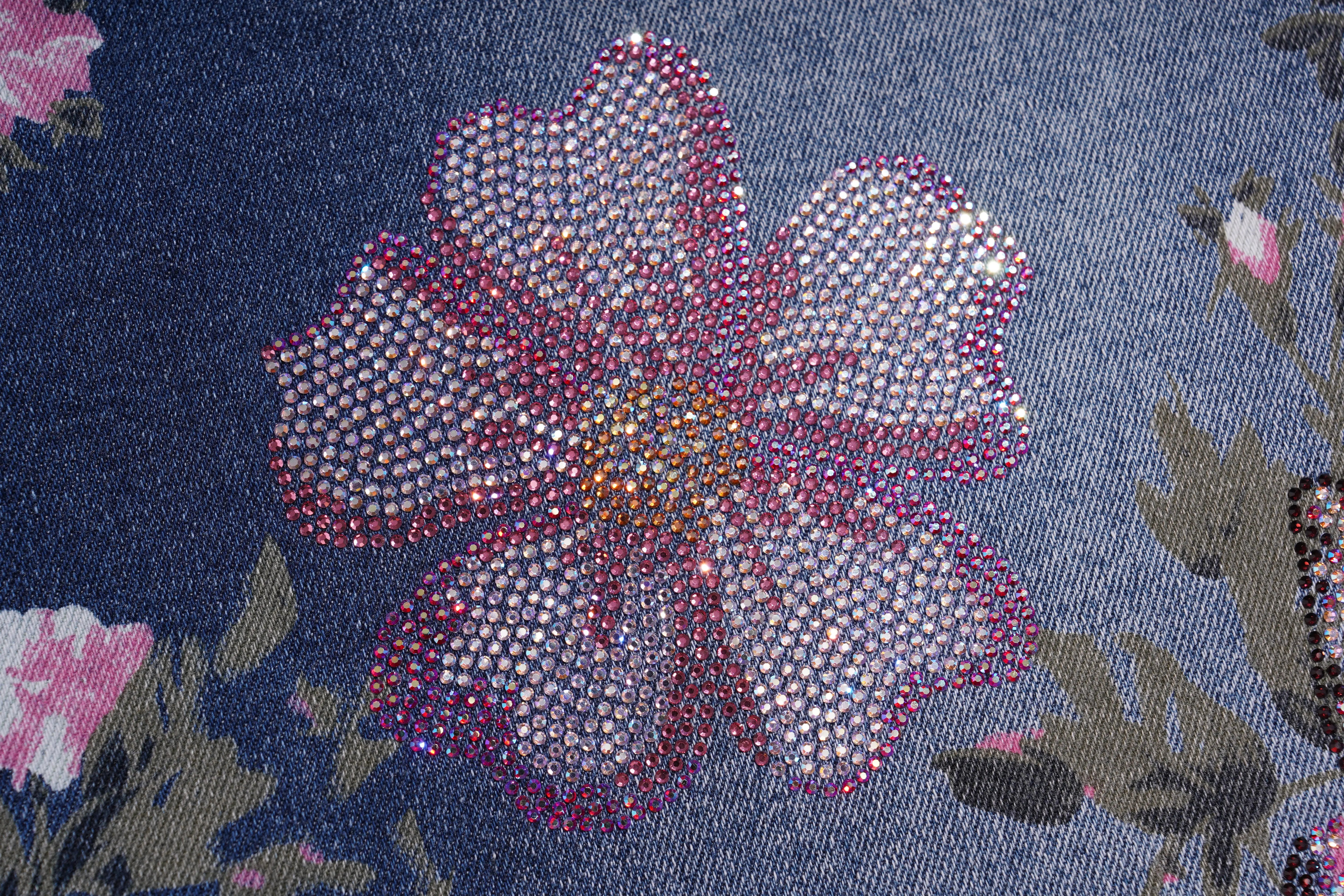 A close-up of a floral embellishment on a dark blue fabric background. The flower is made of many small rhinestones, with pink and silver tones, and is surrounded by matching floral details on a textured blue textile. TCH Floral Butterfly Print Denim Midi Skirt