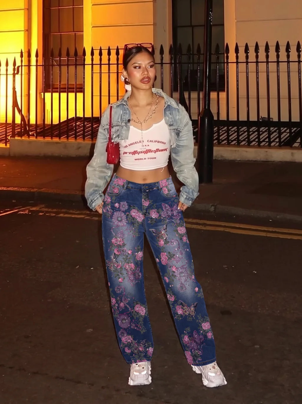 A woman stands on a city street at night wearing a light-wash denim cropped jacket over a white graphic crop top, paired with blue floral wide-leg jeans and white sneakers. She is holding a small red handbag. TCH Floral Butterfly Rhinestone Wide-Leg Denim Jeans