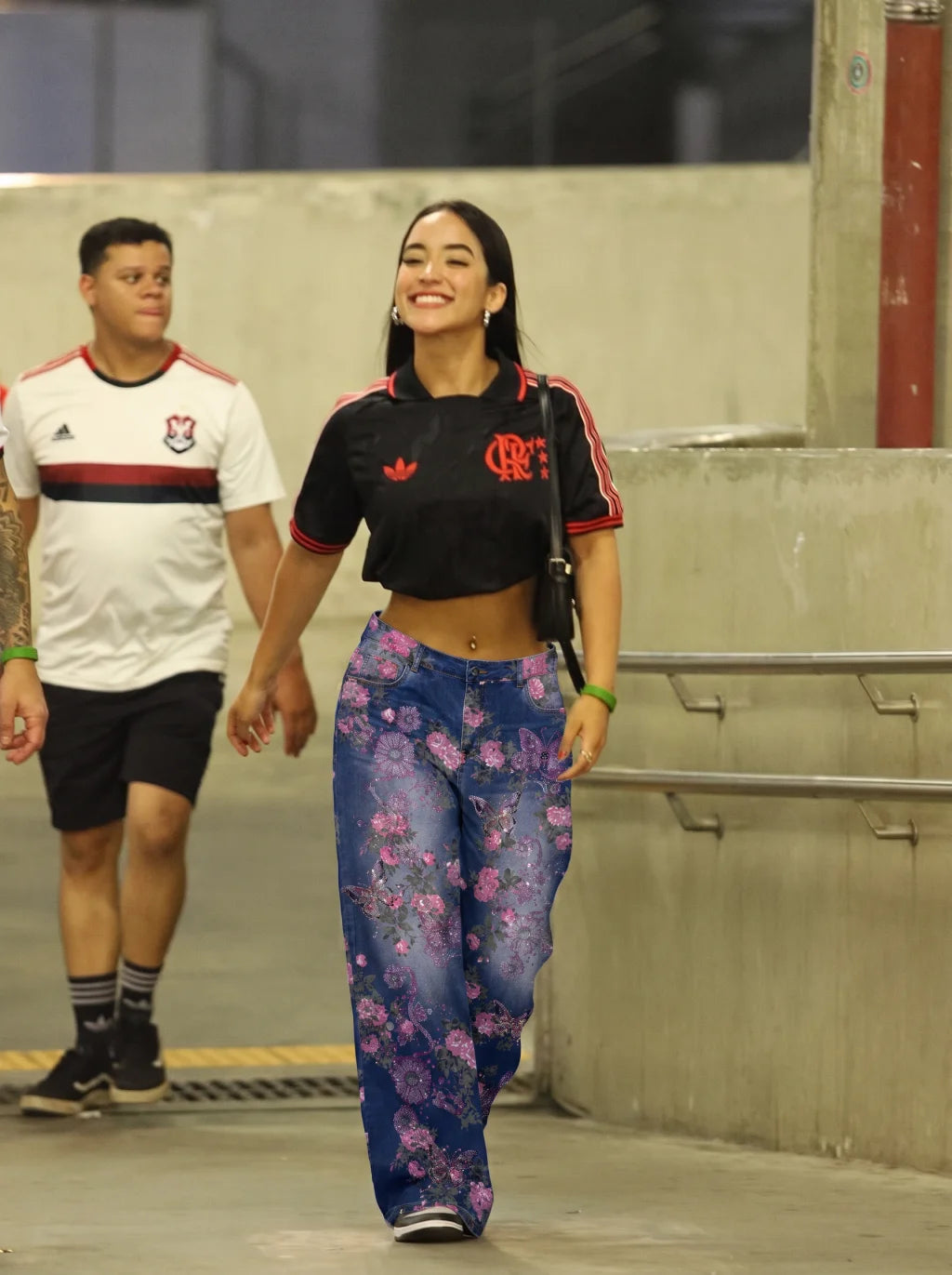 A woman walking in an indoor corridor wearing a black cropped Adidas polo shirt with red details and wide-leg blue jeans with a pink floral pattern. A man in a white Flamengo jersey walks behind her. TCH Floral Butterfly Rhinestone Wide-Leg Denim Jeans