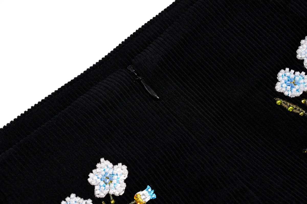 Close-up of a black ribbed corduroy garment, likely a skirt or shorts, featuring embroidered white, light blue, and yellow floral details along the hem. TCH Floral Embroidered Techno Corduroy Mini Skirt