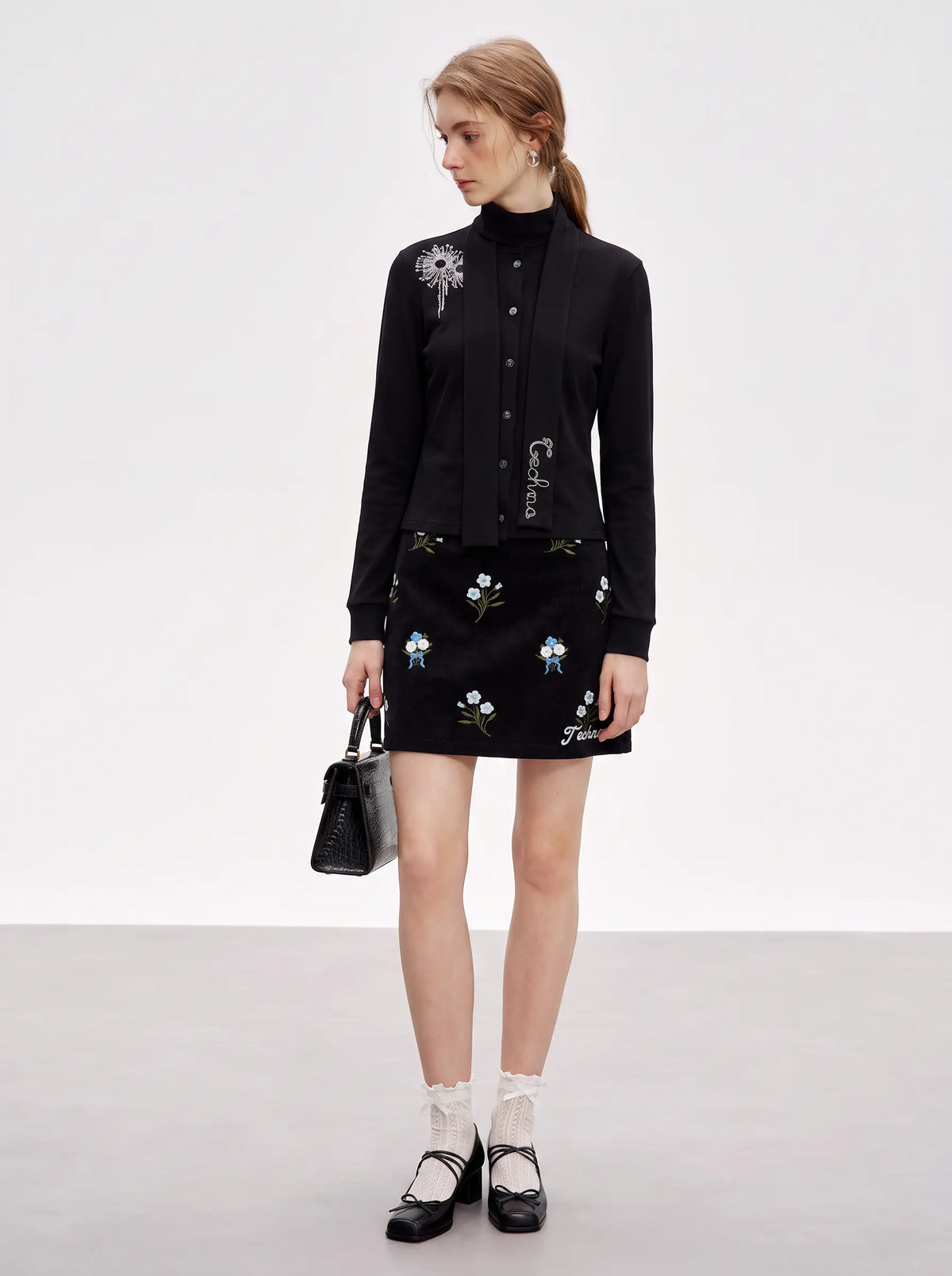 A model stands in a studio wearing a black long-sleeve button-front top and matching black mini skirt, both decorated with embroidered floral and butterfly motifs. She is holding a small black handbag and wearing white ruffled socks with black Mary Jane shoes. TCH Floral Embroidered Techno Corduroy Mini Skirt