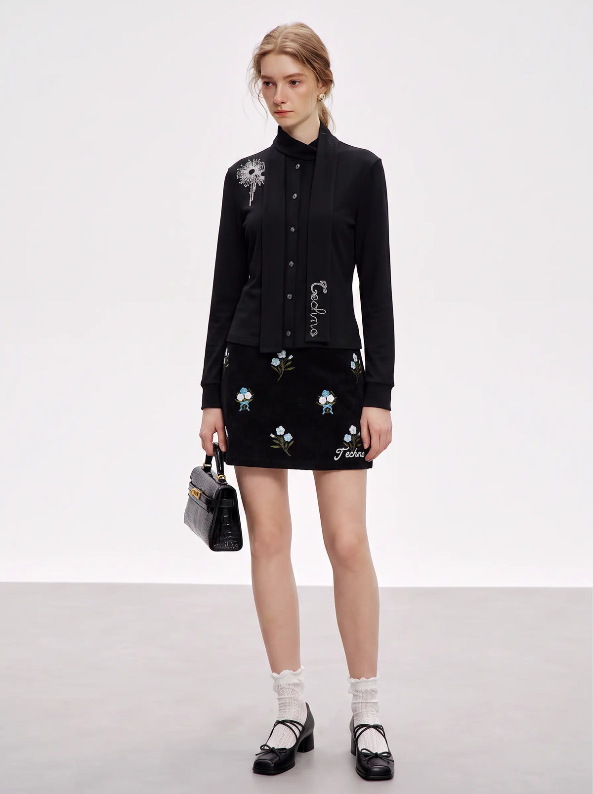 A model stands in a studio wearing a black long-sleeve button-front jacket with floral embroidery and a matching black mini skirt with embroidered flowers. She is holding a small black handbag and wearing white socks and black Mary Jane shoes. TCH Floral Embroidered Techno Corduroy Mini Skirt