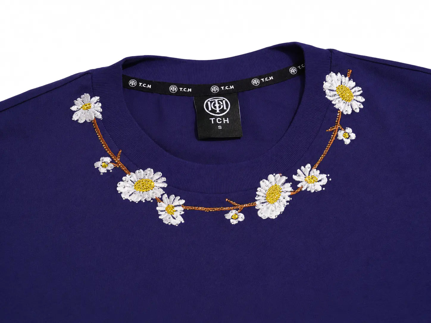 Close-up of a royal blue crewneck sweatshirt featuring a decorative embroidered daisy garland across the collar. The neck label shows the brand 'TCH' (Tchic). TCH Floral Embroidery Graphic Navy Blue T-Shirt