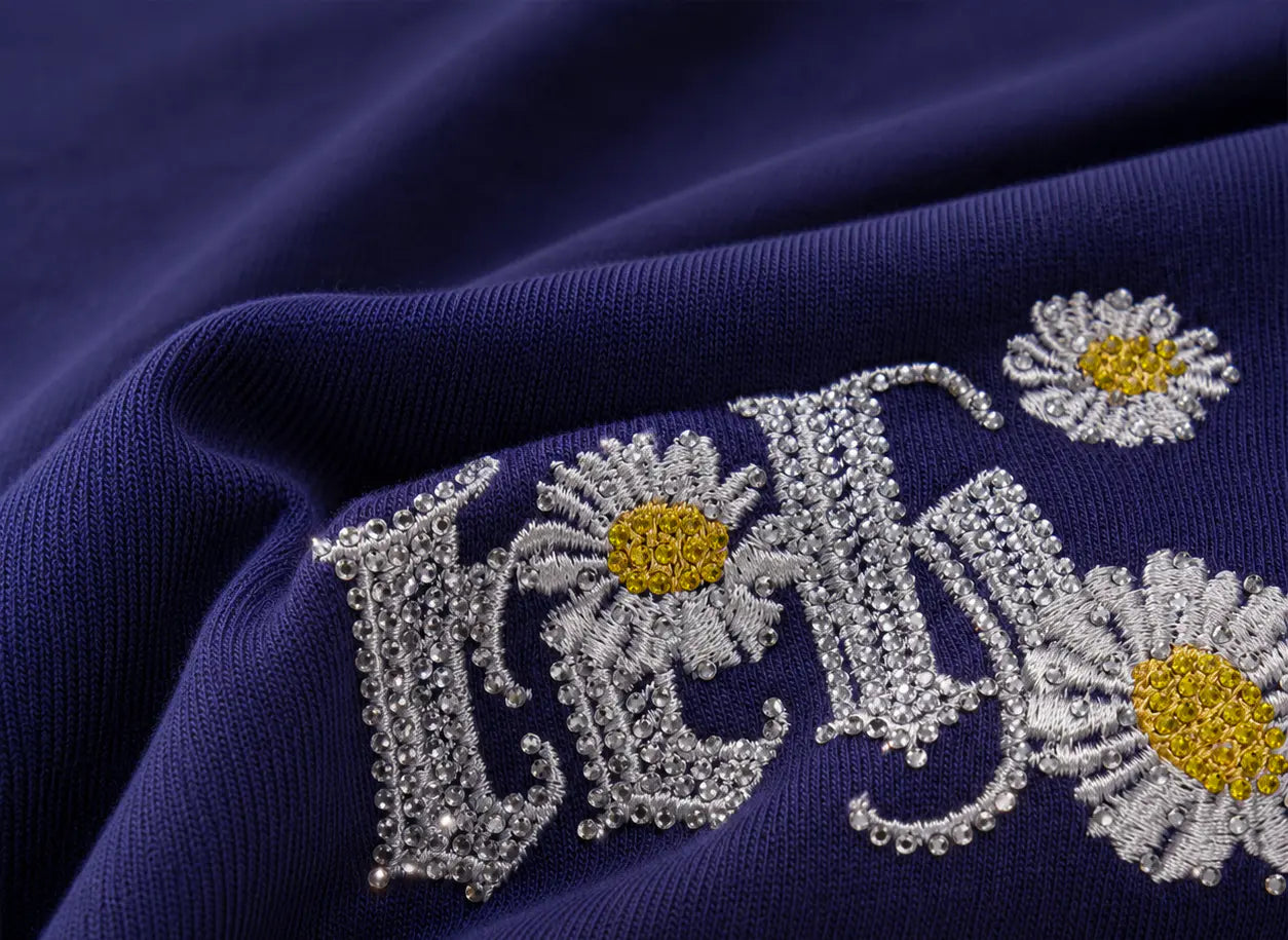 Close-up of a navy blue knit fabric featuring decorative embroidered lettering and daisy flowers with silver beads and yellow centers. TCH Floral Embroidery Graphic Navy Blue T-Shirt