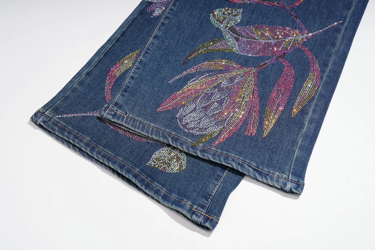 A close-up of a folded pair of blue denim jeans featuring large, colorful floral embellishments made from sequins and rhinestones in pink, purple, gold, and green on the leg. TCH Floral Print Wide-Leg Denim Jeans