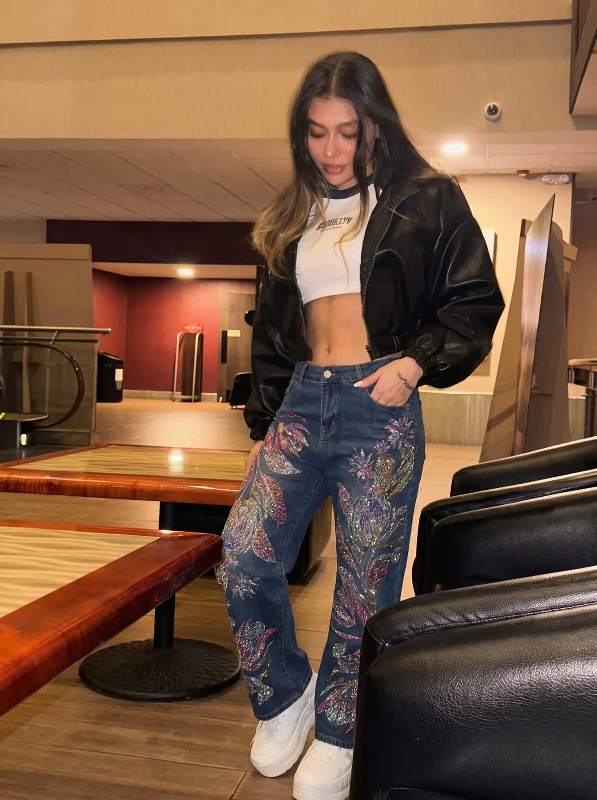 A woman posing in a stylish outfit featuring blue jeans with colorful paisley embroidery, a black leather jacket, a white crop top, and white sneakers, set in a modern indoor environment. TCH Floral Print Wide-Leg Denim Jeans