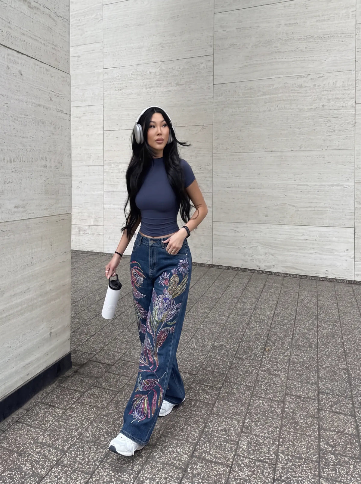 A woman walking outdoors in a modern concrete corridor, wearing a dark blue ribbed top, blue jeans with colorful butterfly wing embroidery on the lower legs, white sneakers, and white headphones while holding a white water bottle. TCH Floral Print Wide-Leg Denim Jeans