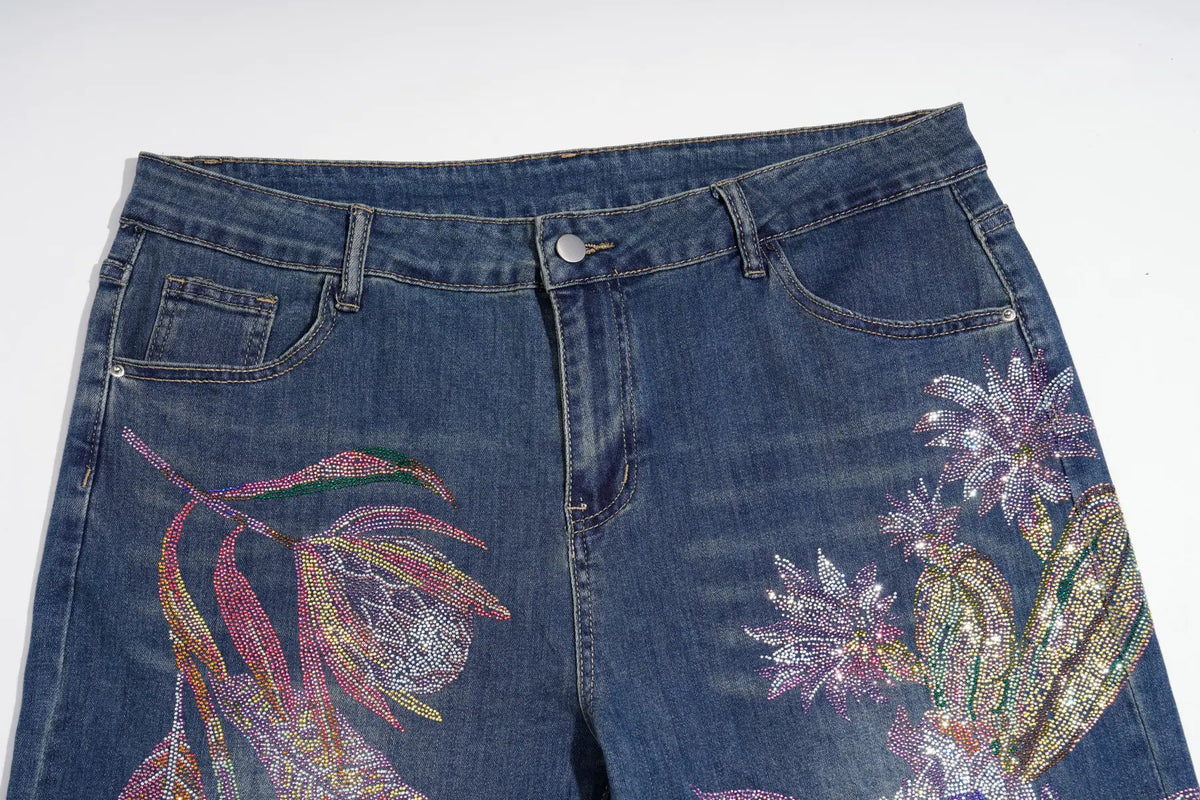 Close-up of blue denim jeans with large, colorful floral and botanical embellishments on the front legs, featuring pink, purple, green, and gold sequins or rhinestones on a white background. TCH Floral Print Wide-Leg Denim Jeans