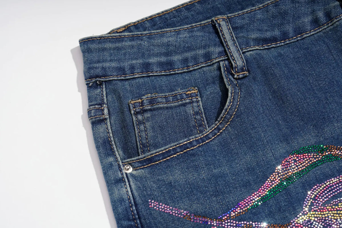 Close-up of blue denim jeans showing the back pocket, belt loop, and a colorful rhinestone embellishment on the lower right leg against a white background. TCH Floral Print Wide-Leg Denim Jeans