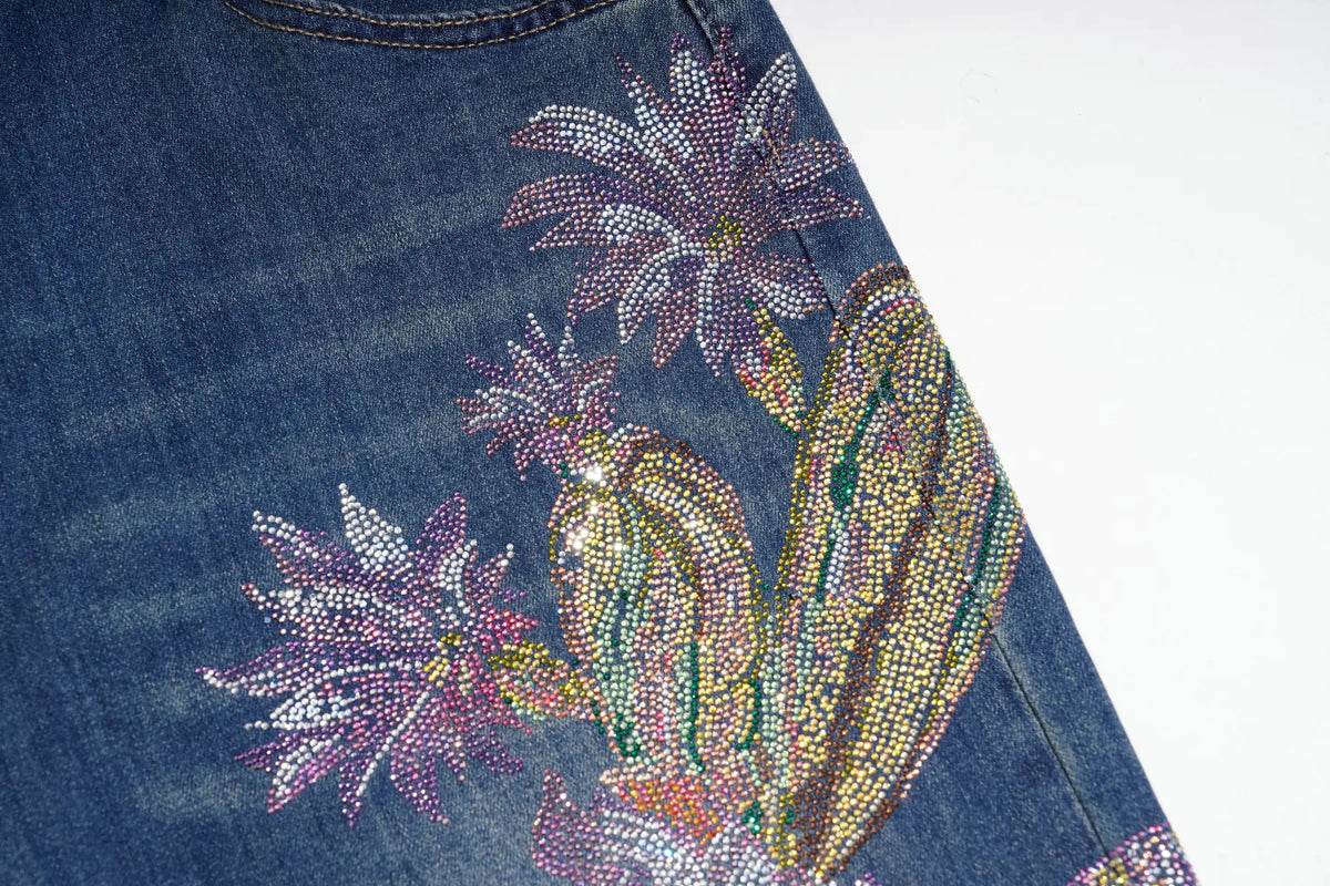 Close-up of a blue denim garment featuring a large rhinestone-embellished floral and paisley design in pink, purple, green, and gold tones on a white background. TCH Floral Print Wide-Leg Denim Jeans