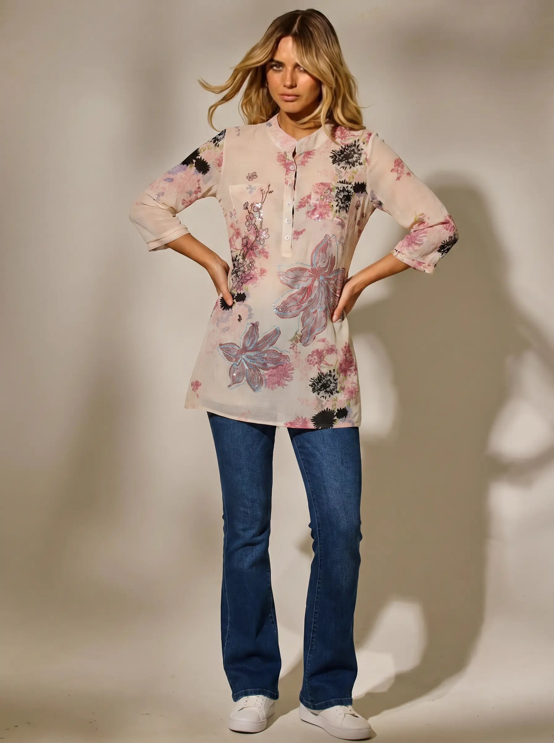 A woman modeling a sheer, light pink floral tunic top with blue jeans and white sneakers, standing against a warm beige studio backdrop. TCH Floral Rhinestone Butterfly Print Short Sleeve Blouse