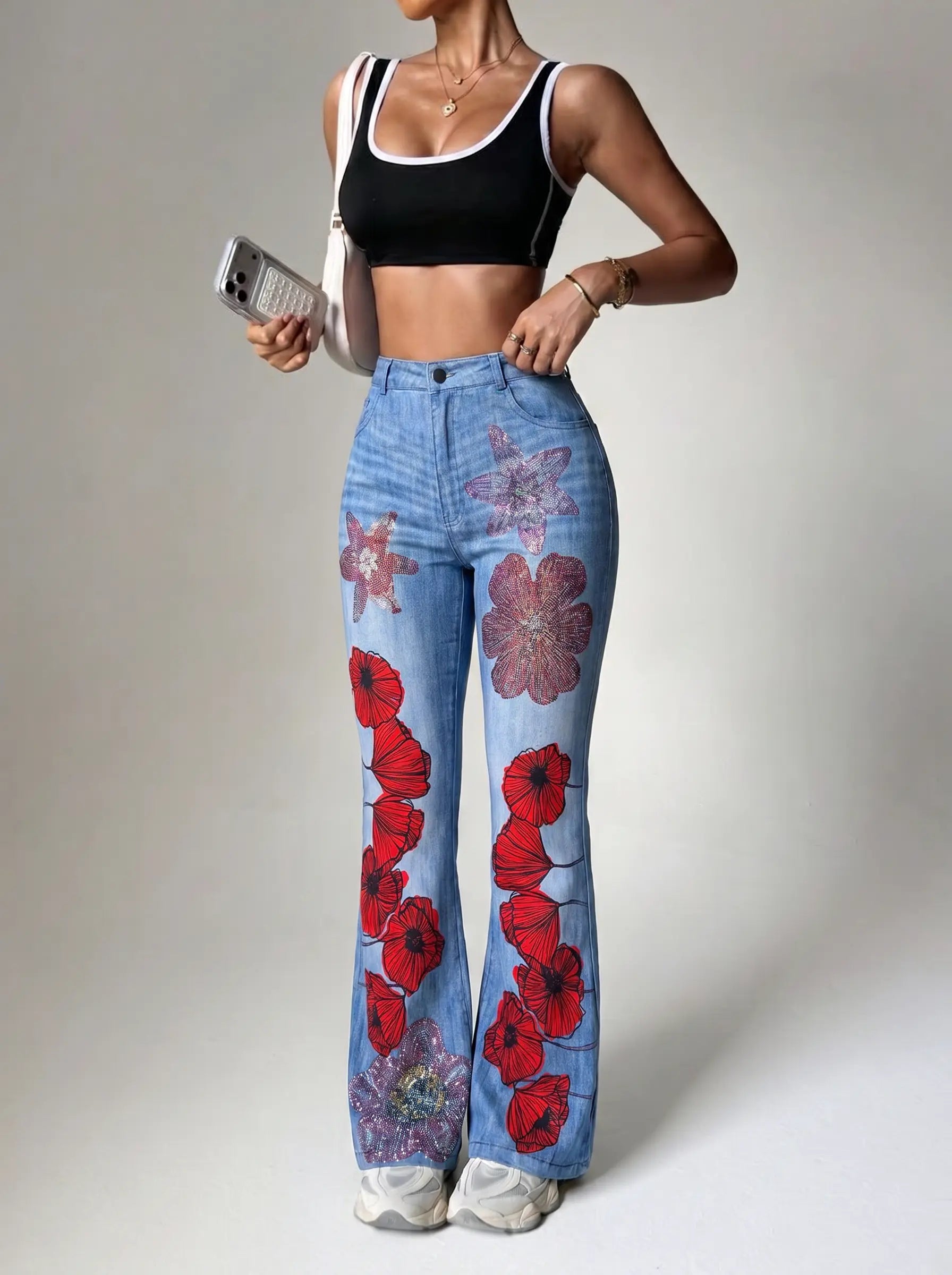 A model wearing high-waisted, wide-leg blue jeans with a red poppy and purple floral print, paired with a black sports bra and white sneakers, holding a silver smartphone against a neutral studio background. TCH Floral Rhinestone Wide Leg Denim Jeans