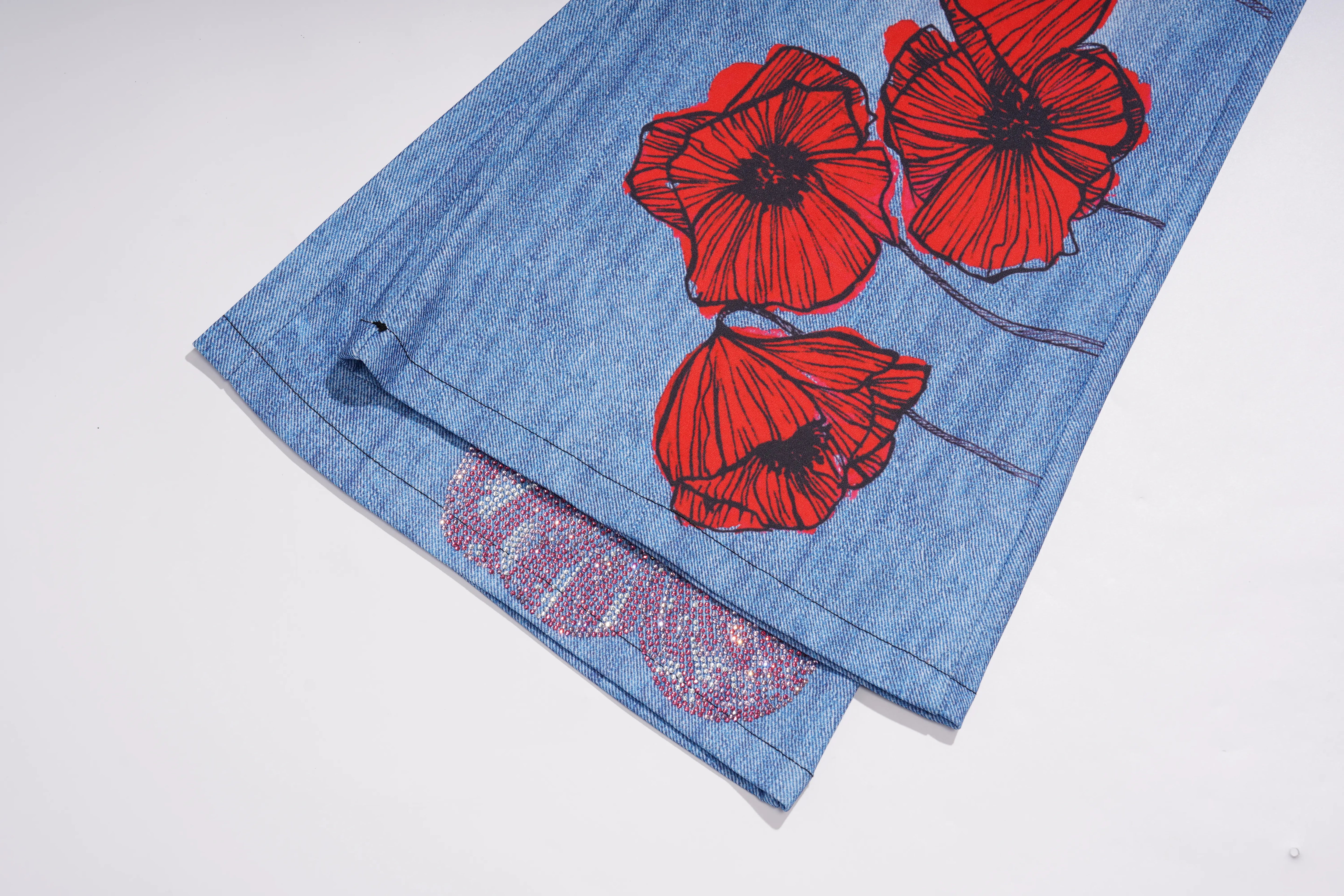 A close-up of a light blue denim garment, likely a skirt or shorts, featuring a graphic print of three red poppies with black centers and a pink lace applique near the hem, shown on a plain white background. TCH Floral Rhinestone Wide Leg Denim Jeans