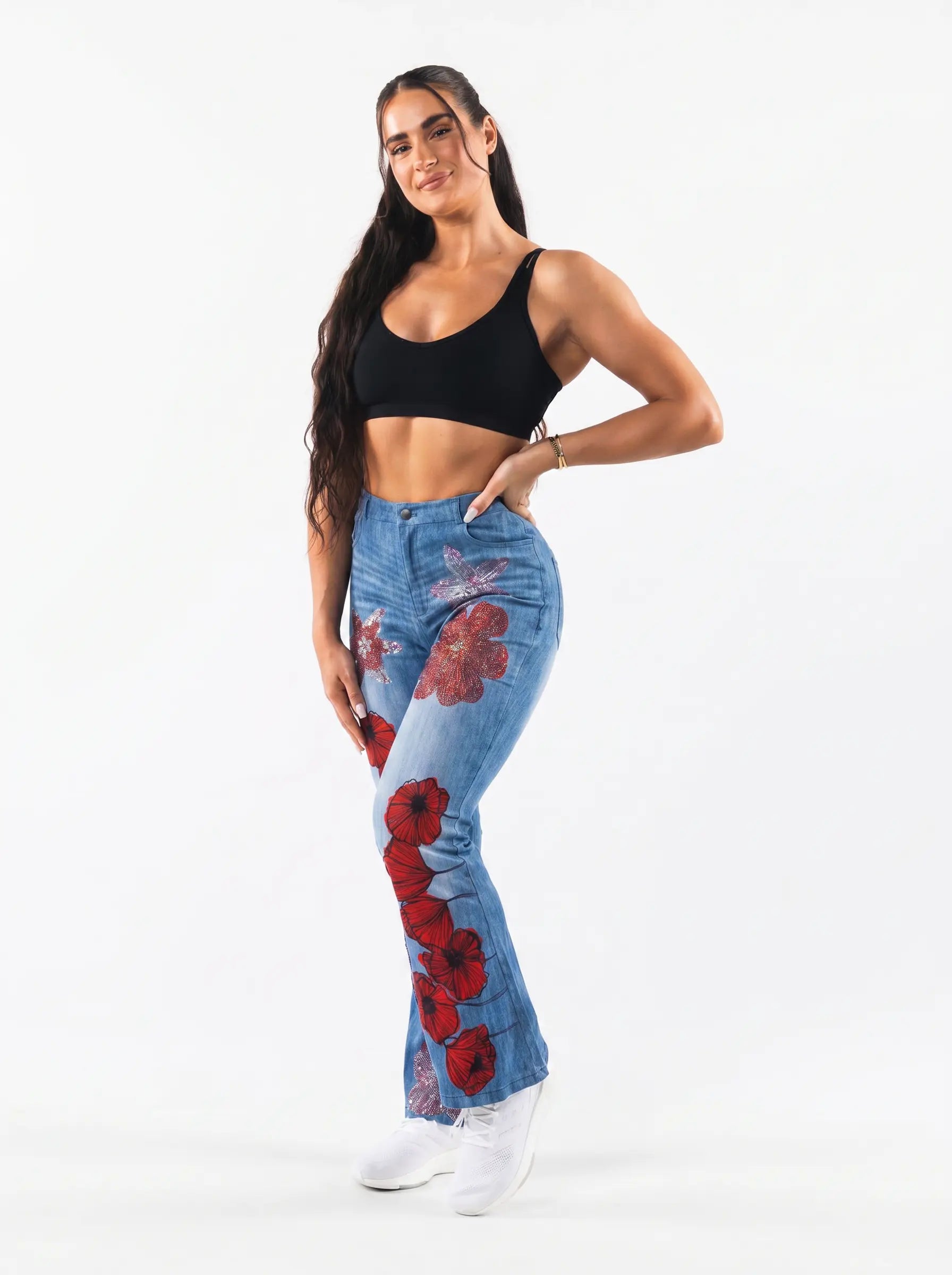 A woman modeling a pair of light blue denim flare jeans with red poppy floral prints, styled with a black crop top and white sneakers against a plain white studio background. TCH Floral Rhinestone Wide Leg Denim Jeans