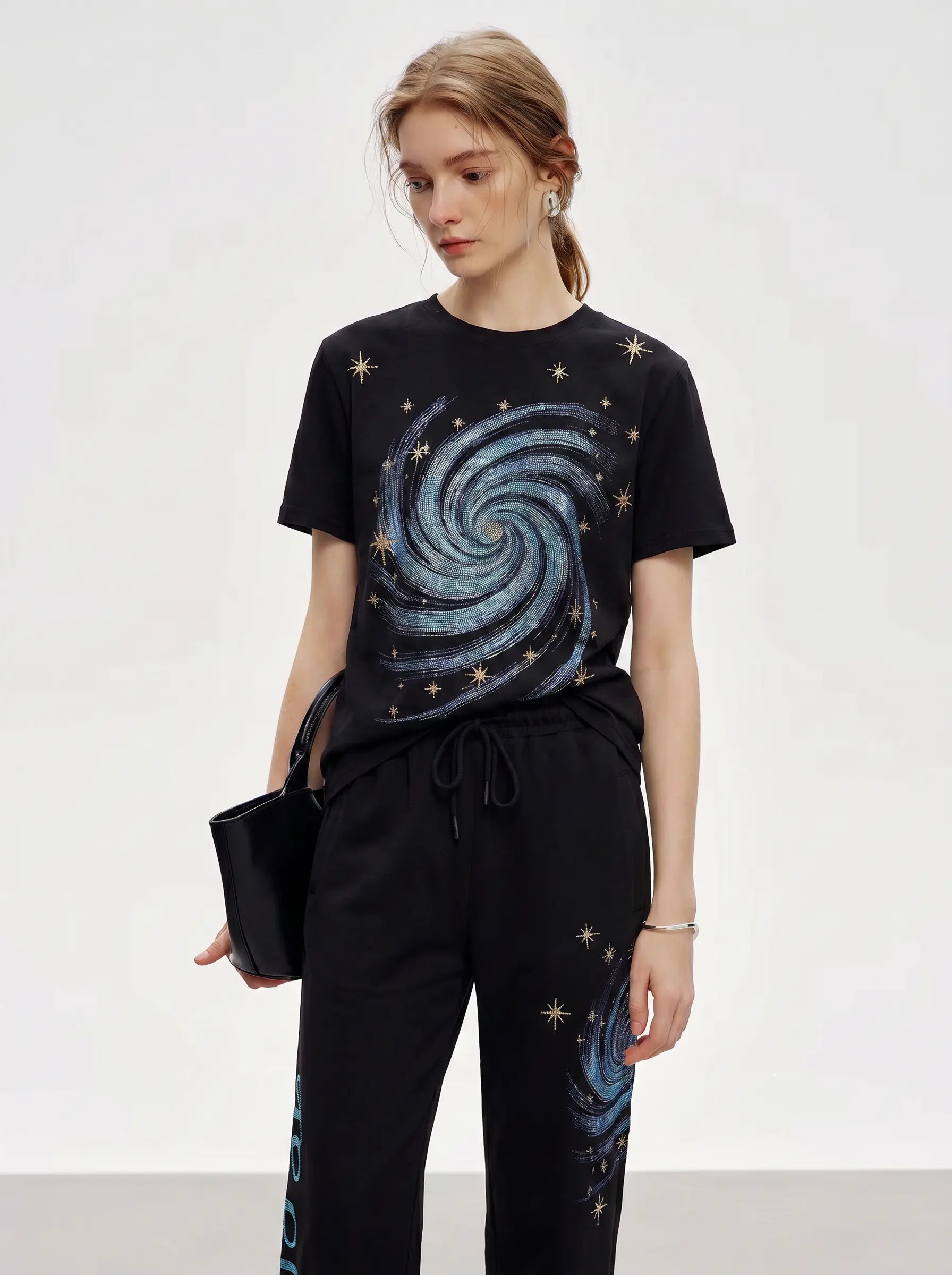 A woman modeling a black short-sleeve t-shirt with a blue and silver spiral galaxy print and matching black sweatpants with a galaxy print on the leg, holding a black clutch bag against a light gray studio background. TCH Galaxy Rhinestone Wide Leg Sweatpants