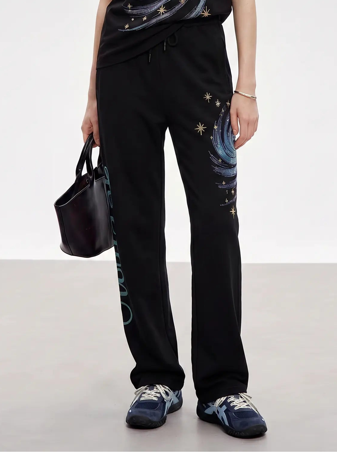 A model stands in a studio wearing a black t-shirt and matching sweatpants with a galaxy swirl print, holding a small black handbag. TCH Galaxy Rhinestone Wide Leg Sweatpants