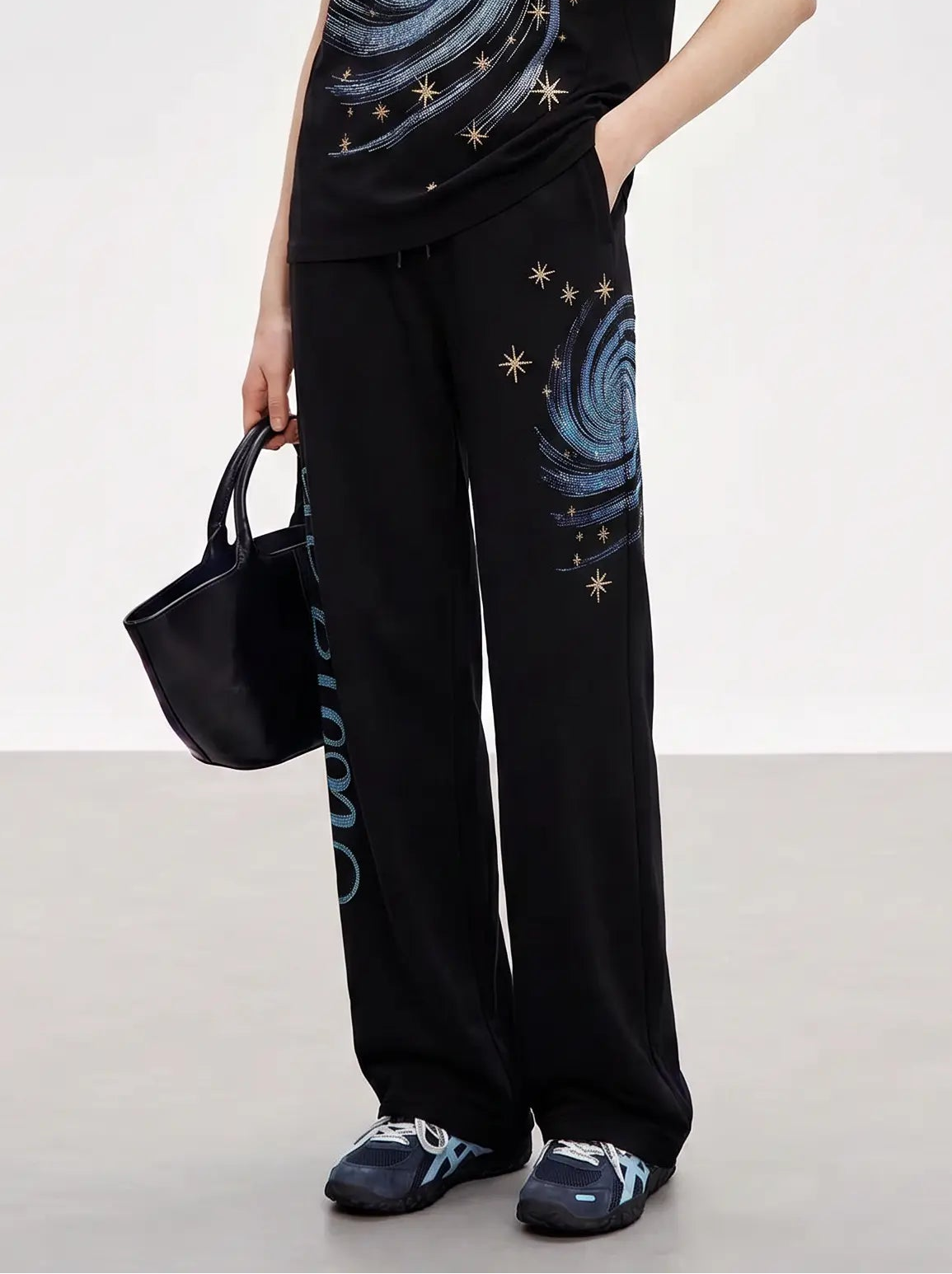 A woman modeling a black short-sleeve t-shirt and matching wide-leg pants set, both featuring a blue spiral galaxy print with small gold star details. She is holding a black handbag and wearing dark blue sneakers, set against a plain light gray studio background. TCH Galaxy Rhinestone Wide Leg Sweatpants