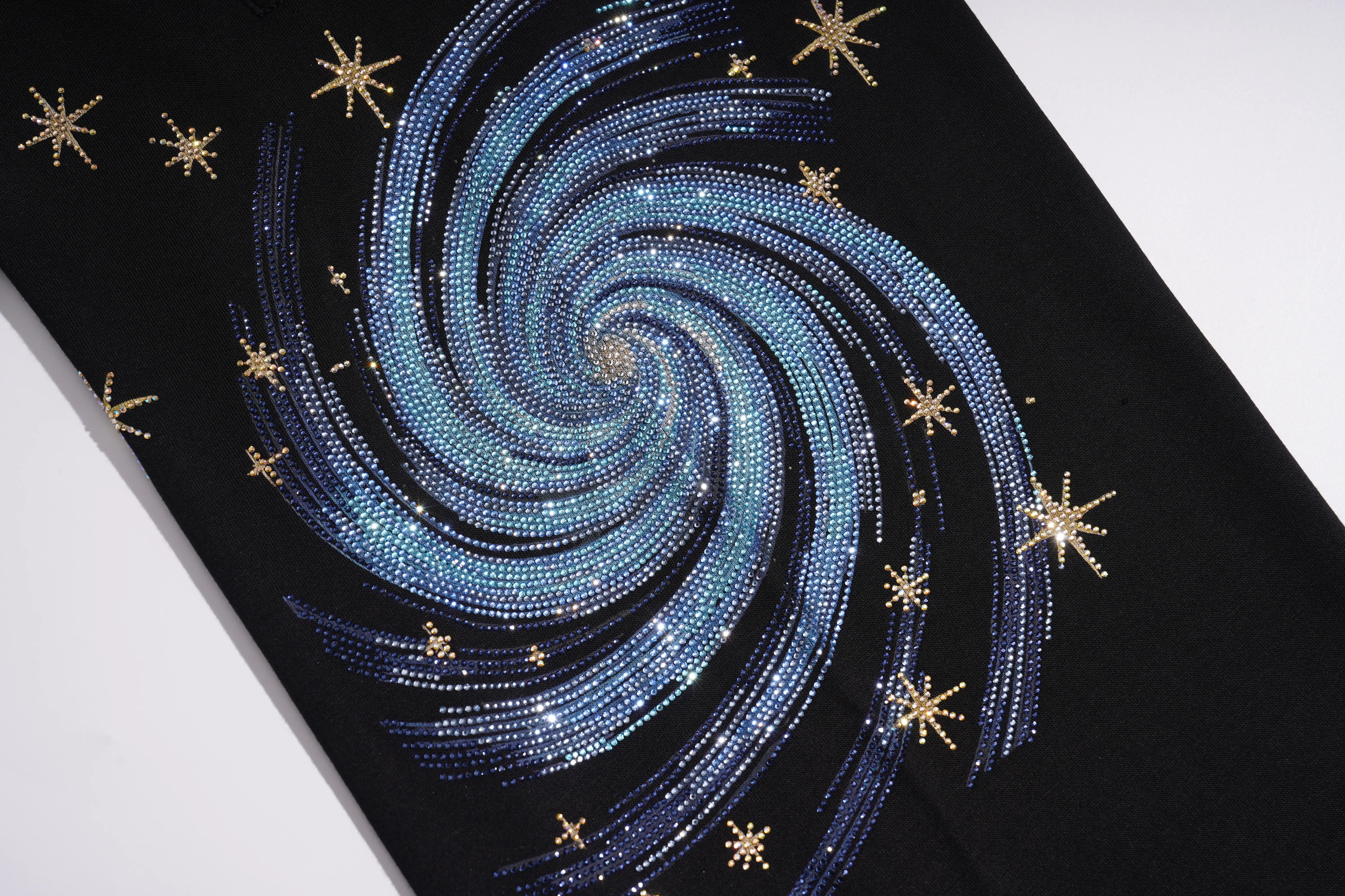 A close-up of a black garment featuring a large spiral galaxy design made of blue and silver rhinestones, accented with small gold star embellishments. TCH Galaxy Rhinestone Wide Leg Sweatpants