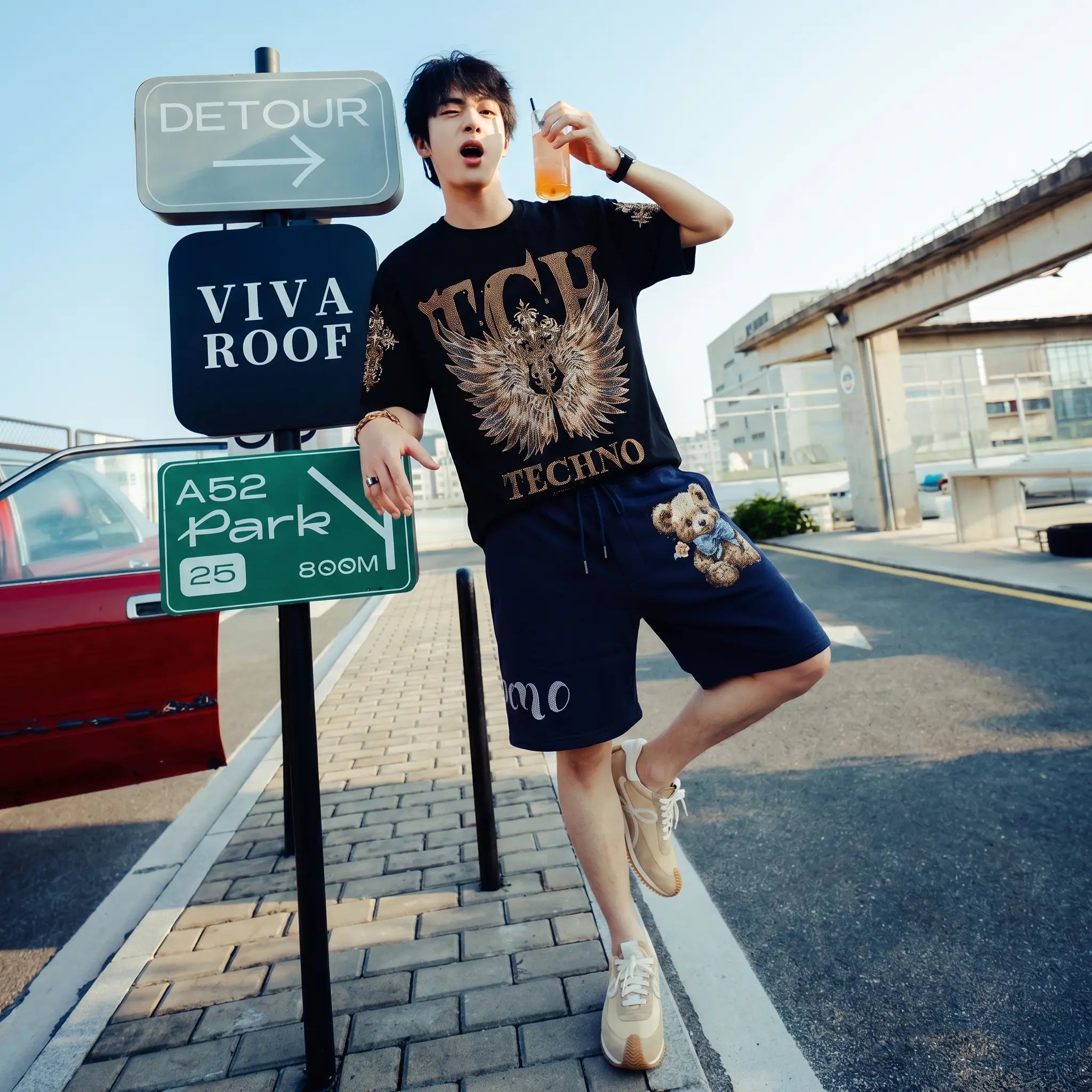 A man posing outdoors in a black graphic t-shirt and navy blue shorts with cartoon bear embroidery, standing next to a road sign post. TCH Golden Wing Techno Oversized Graphic T-Shirt