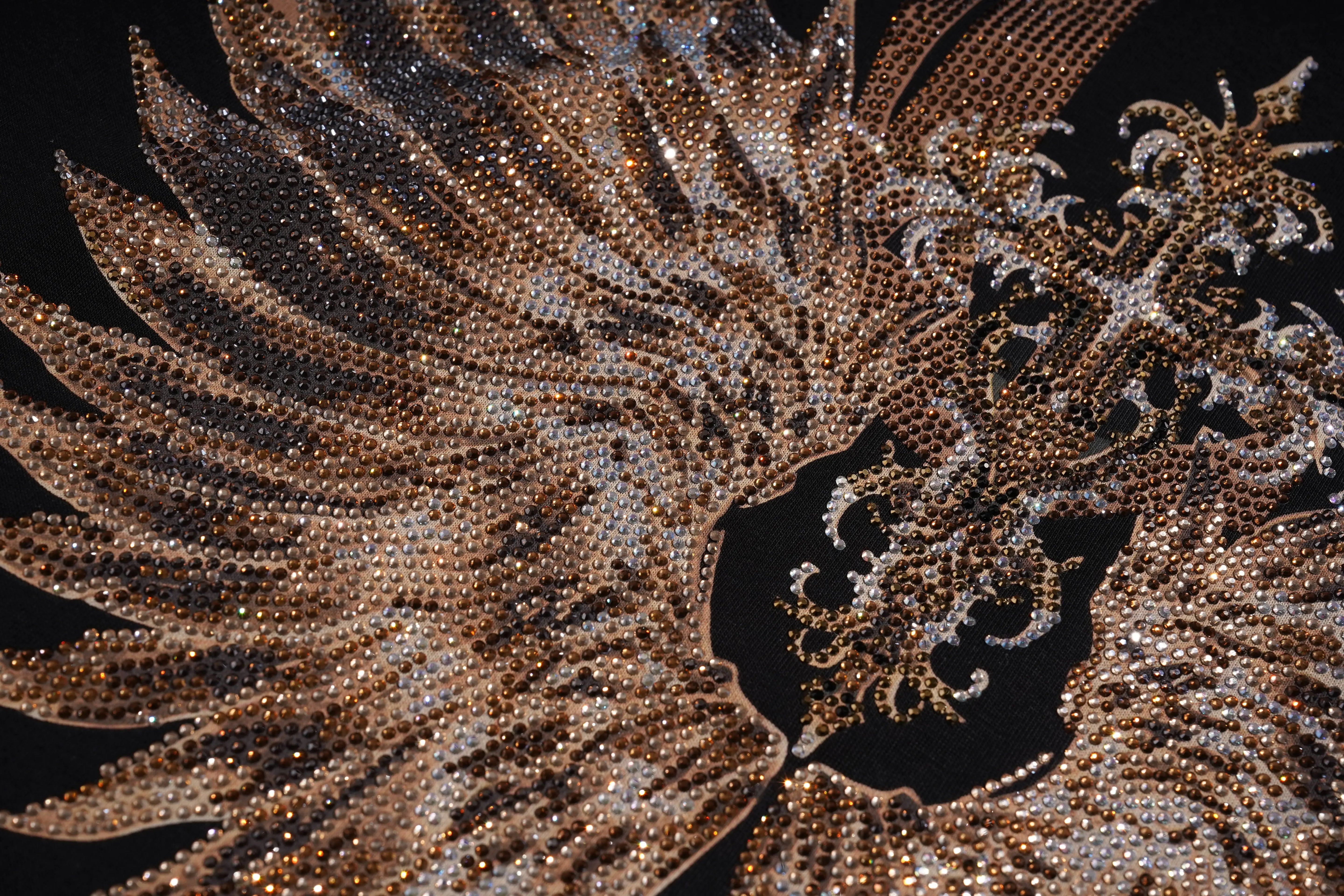 A close-up of an ornate, highly embellished garment or textile featuring intricate beadwork and sequins in metallic bronze, gold, and silver tones against a black background. TCH Golden Wing Techno Oversized Graphic T-Shirt