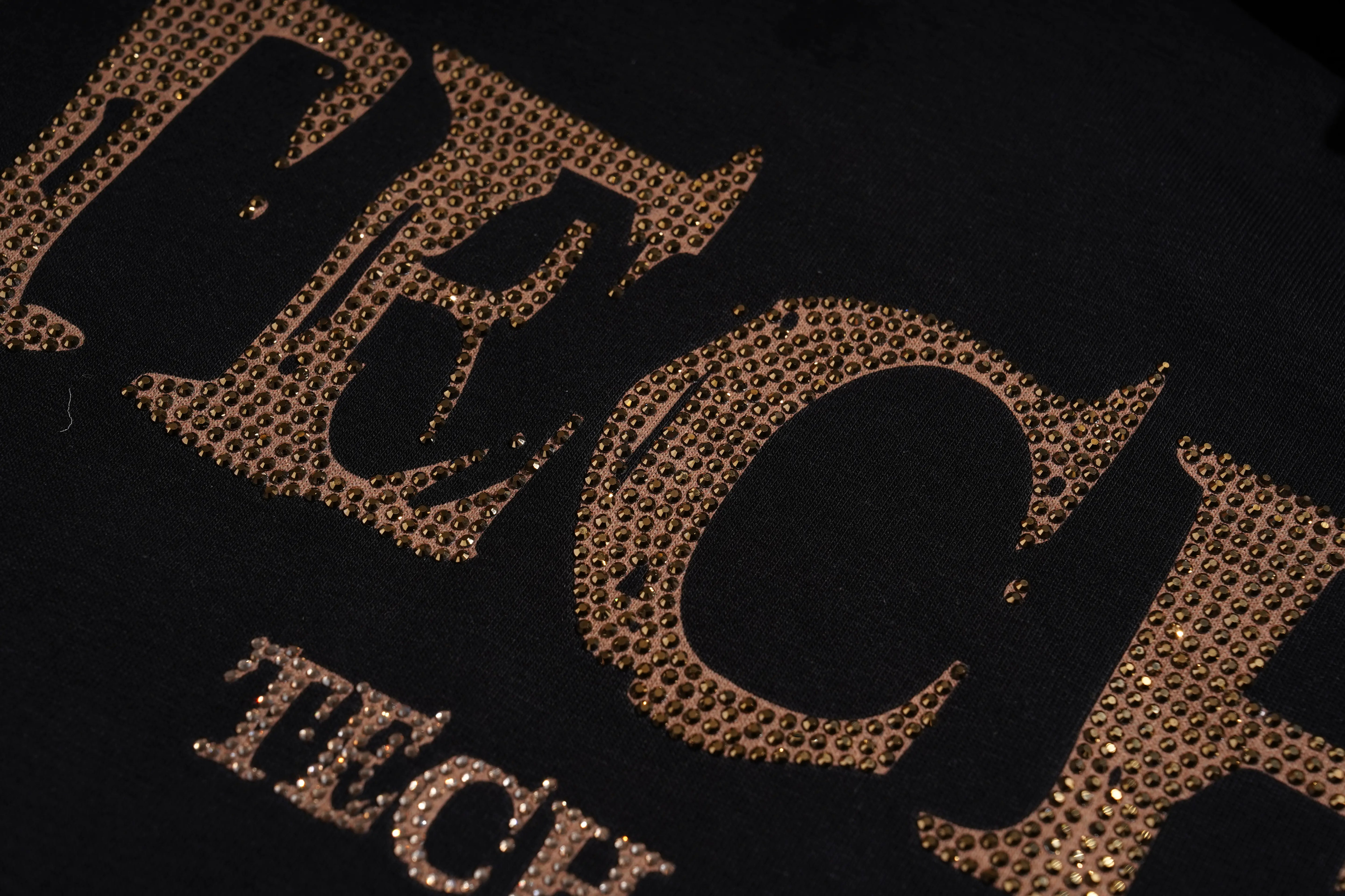 Close-up of a black garment featuring a large metallic rose gold rhinestone-embellished graphic spelling 'TECH' and smaller matching text below. TCH Golden Wing Techno Oversized Graphic T-Shirt