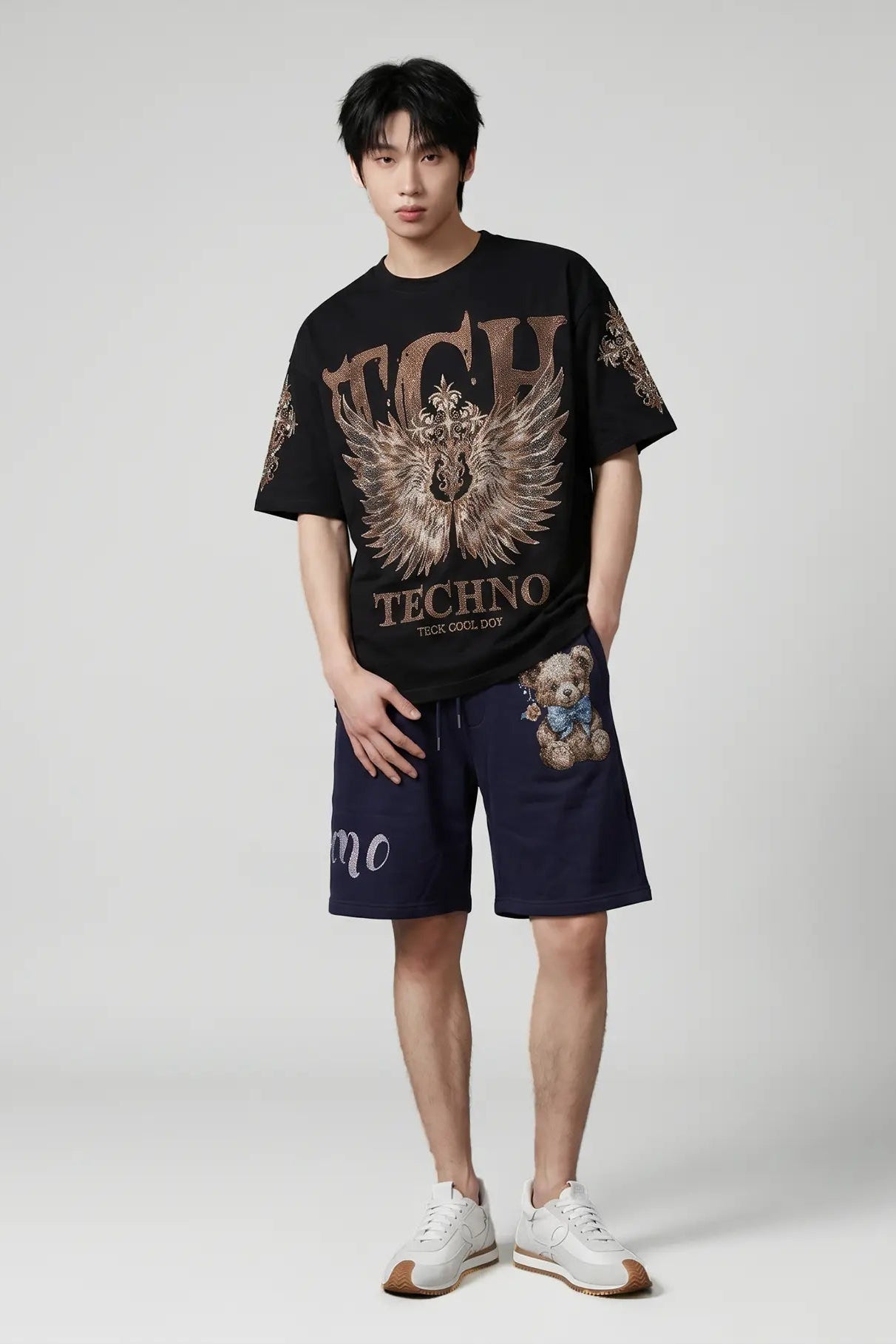 A male model stands against a light gray studio background wearing a black Techno Tour 2024 graphic t-shirt with a large eagle design, navy blue shorts with a teddy bear graphic, and white sneakers. TCH Golden Wing Techno Oversized Graphic T-Shirt