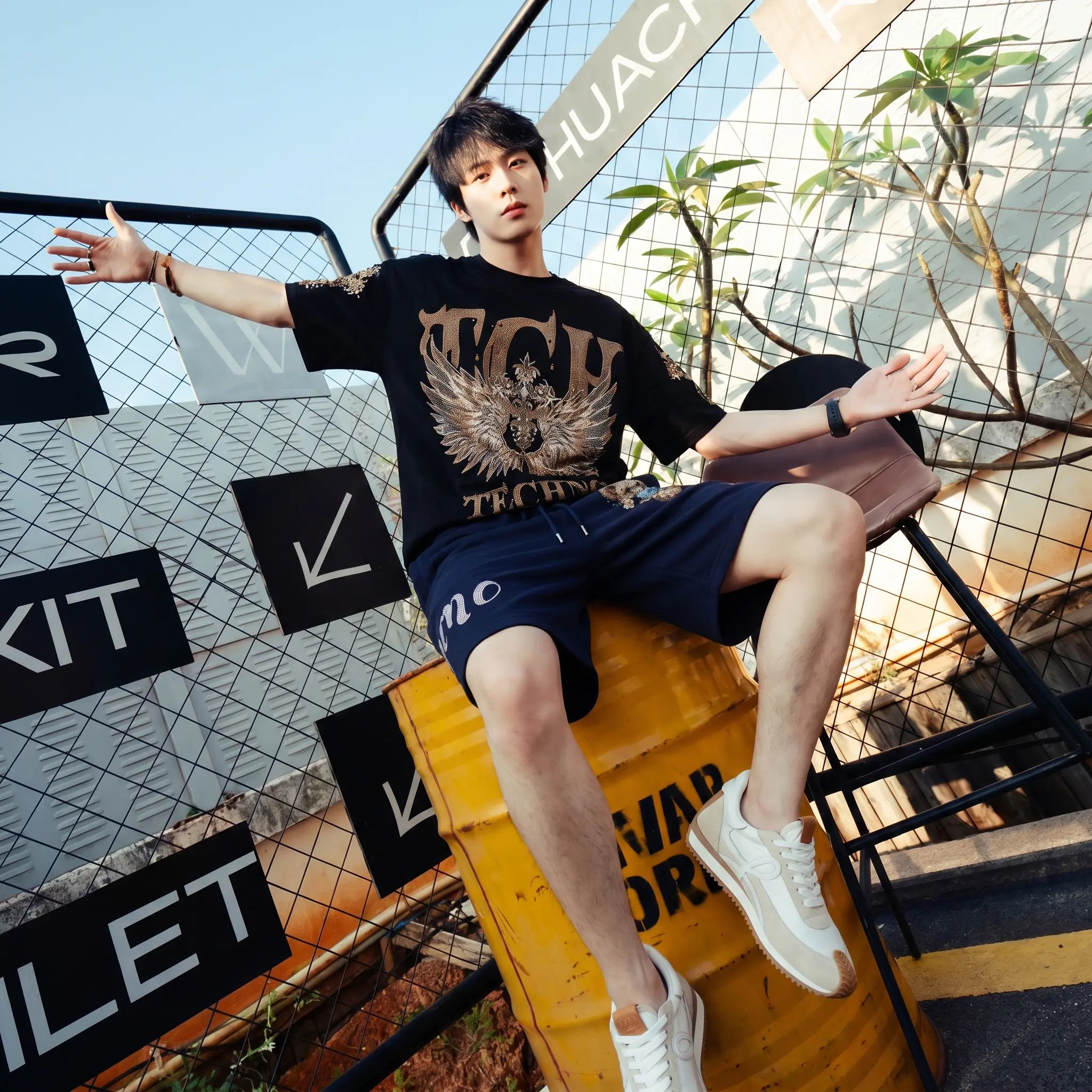 A young man wearing a black graphic t-shirt and navy blue shorts sits on a yellow barrel in front of a metal fence and outdoor background. The t-shirt features a large gold and white eagle graphic with the text 'TCU TROYHUNTER'. TCH Golden Wing Techno Oversized Graphic T-Shirt
