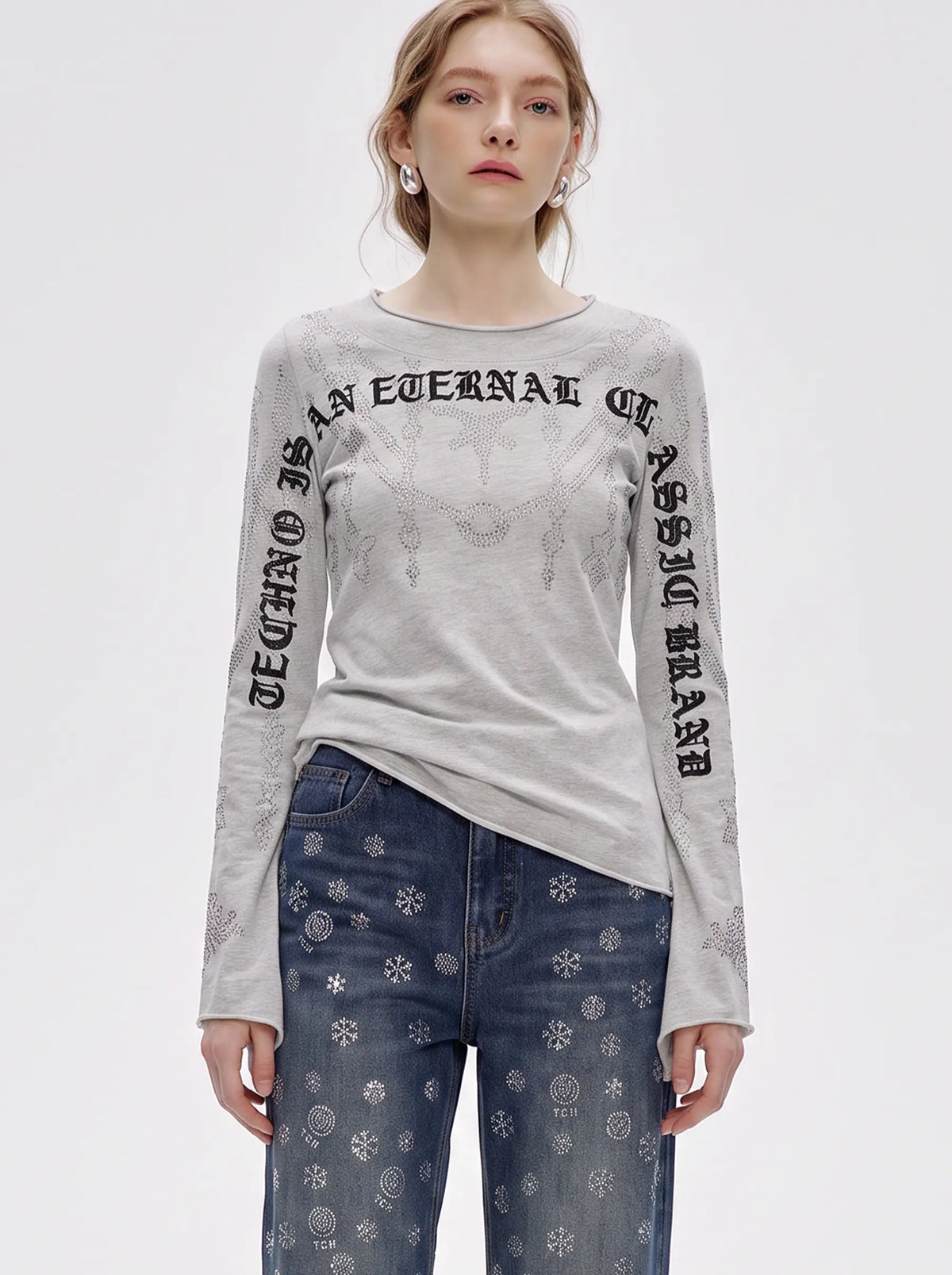 A woman modeling a light gray long-sleeve t-shirt with black gothic-style text printed on the front and sleeves, paired with blue jeans featuring a white snowflake pattern, against a plain light gray studio background. TCH Gothic Rhinestone Lace Pattern Flared Sleeve Long Sleeve Top