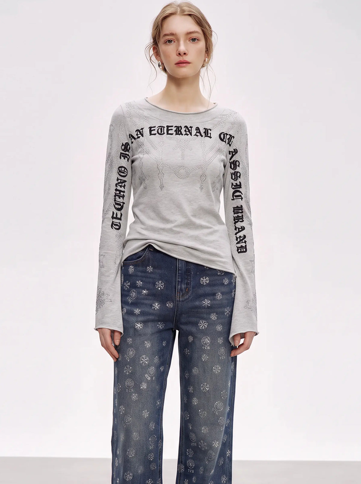 A model wearing a light gray long-sleeve t-shirt with black gothic-style text printed down both sleeves, paired with blue jeans featuring a white snowflake pattern, against a plain white studio background. TCH Gothic Rhinestone Lace Pattern Flared Sleeve Long Sleeve Top