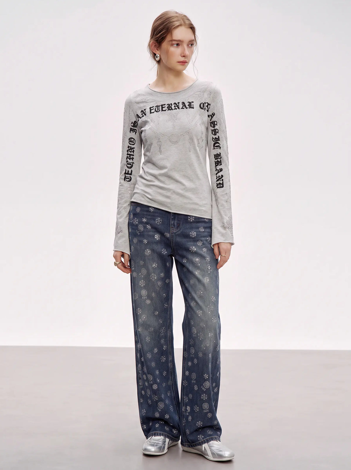A model stands in a studio wearing a light gray long-sleeve top with black text down the sleeves and blue jeans with a small white pattern, paired with silver sneakers. TCH Gothic Rhinestone Lace Pattern Flared Sleeve Long Sleeve Top