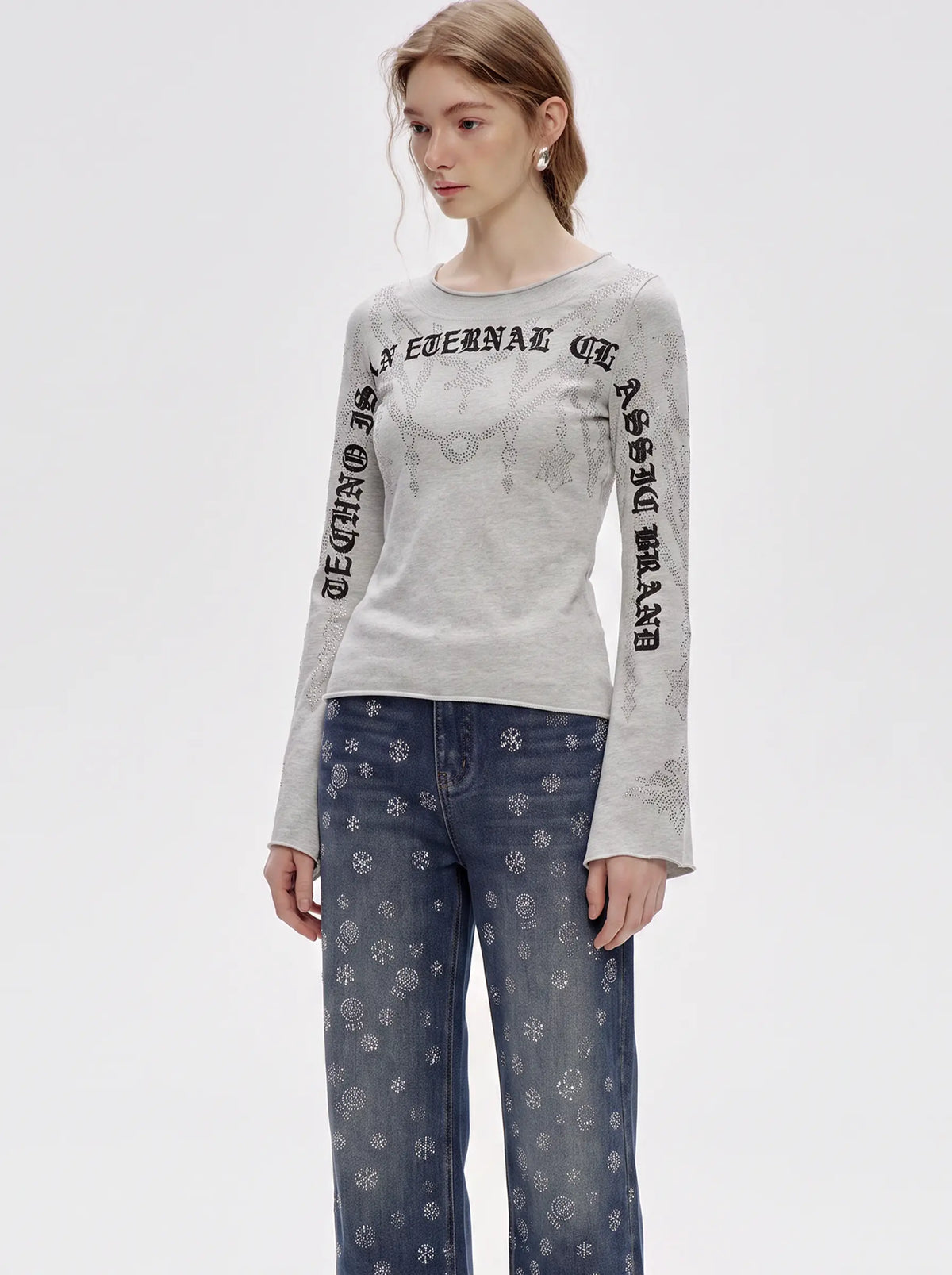 A model wearing a light gray long-sleeve t-shirt with black gothic-style text printed down both sleeves, paired with blue jeans featuring a white snowflake pattern, against a plain light gray background. TCH Gothic Rhinestone Lace Pattern Flared Sleeve Long Sleeve Top