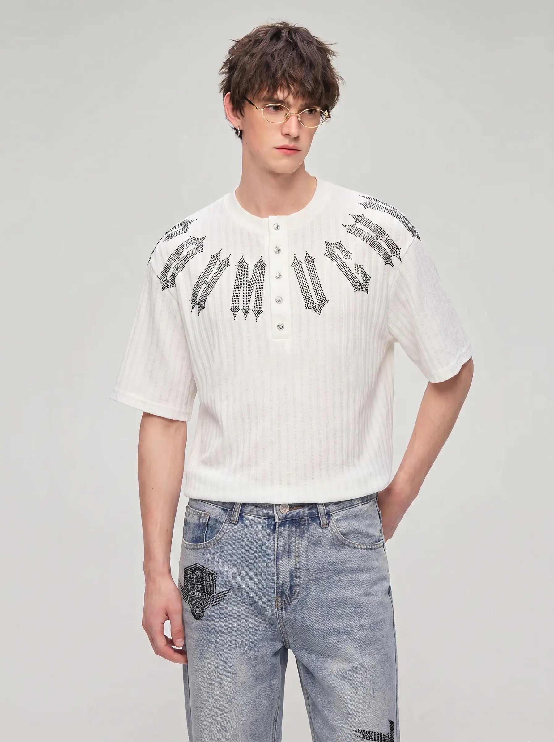 A male model wearing a white short-sleeve ribbed henley shirt with a gray gothic-style graphic across the chest, paired with light-wash blue jeans, against a plain light gray studio background. TCH Gothic Rhinestone Letter Ribbed Henley T-Shirt