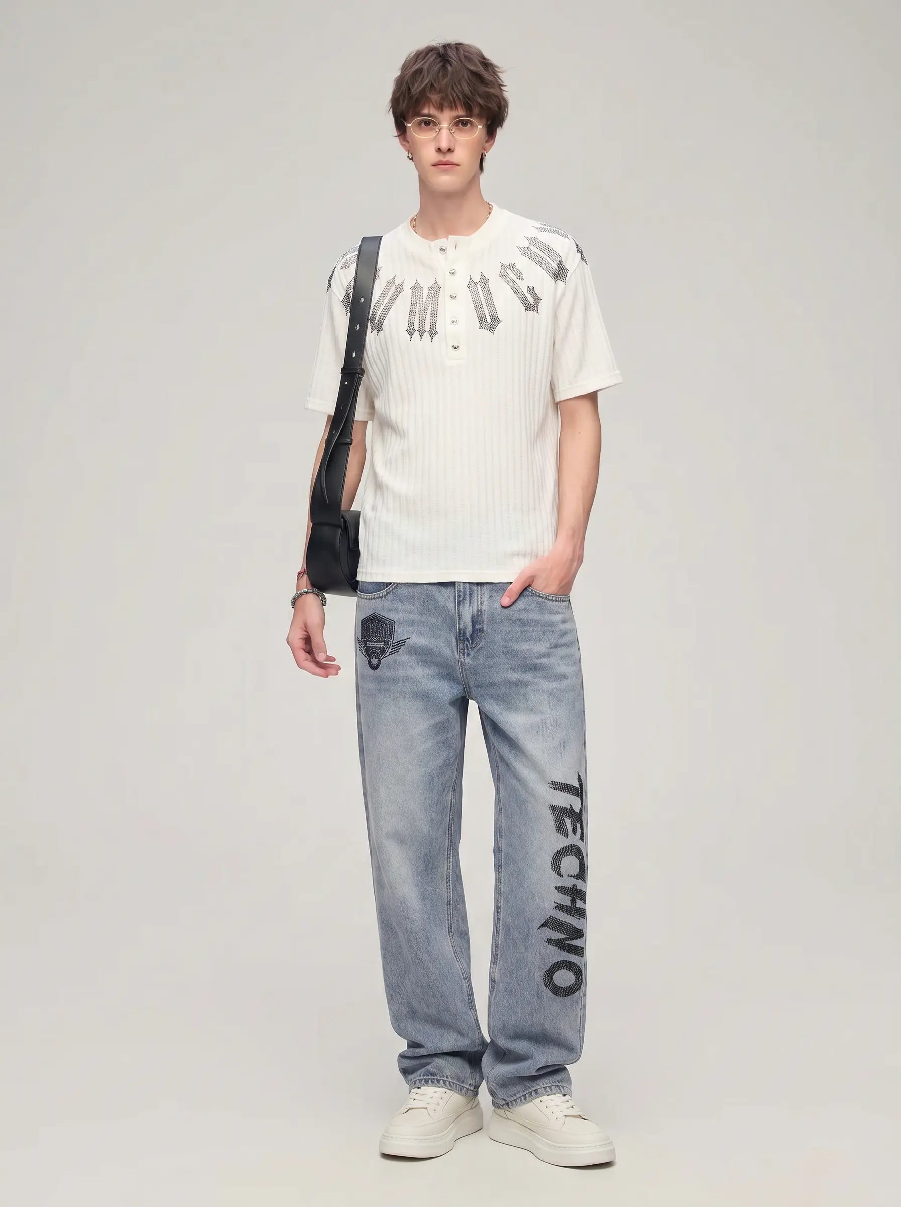 A male model stands against a light gray studio background wearing a white ribbed short-sleeve henley shirt with black graphic lettering, light blue jeans with black text graphics, and white sneakers, while carrying a black shoulder bag. TCH Gothic Rhinestone Letter Ribbed Henley T-Shirt