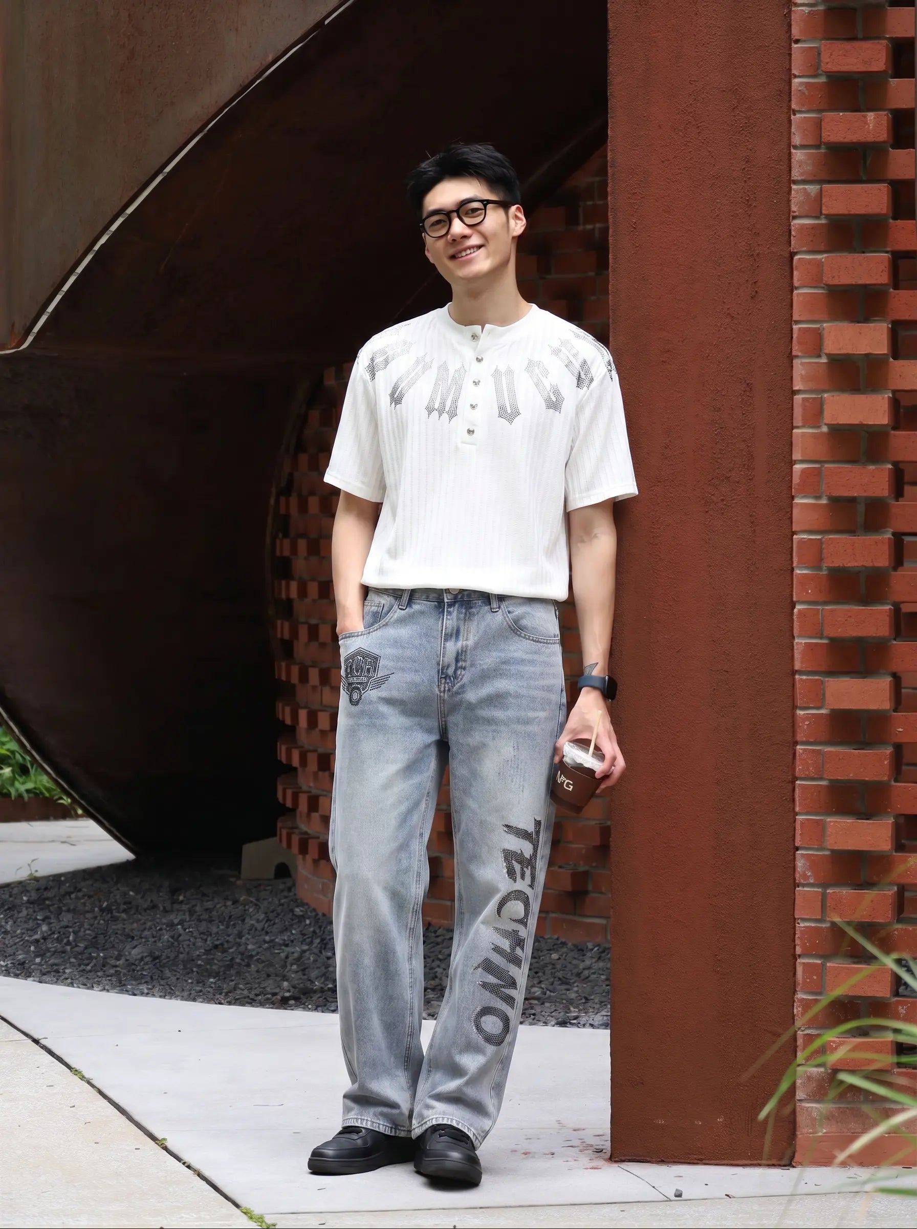 A man wearing a white short-sleeve henley shirt with graphic prints on the chest, light-wash blue jeans with 'TECHNO' printed on the leg, and black shoes, standing outdoors next to a brick column. TCH Gothic Rhinestone Letter Ribbed Henley T-Shirt