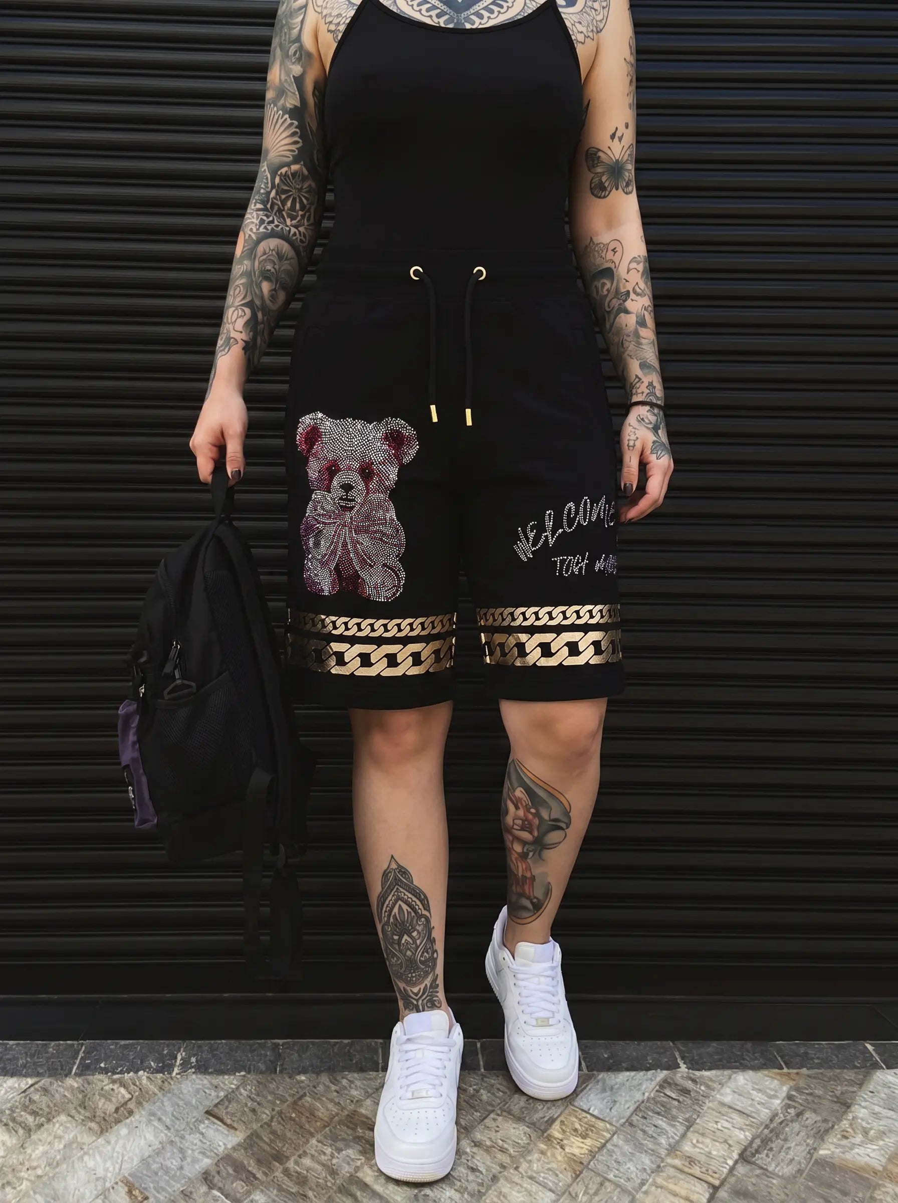 A person wearing a black sleeveless romper with a teddy bear patch on the left leg, a gold chain print hem, white sneakers, and carrying a black backpack, standing in front of a black corrugated metal wall. TCH Gothic Rhinestone Teddy Bear Chain Print Sweat Shorts