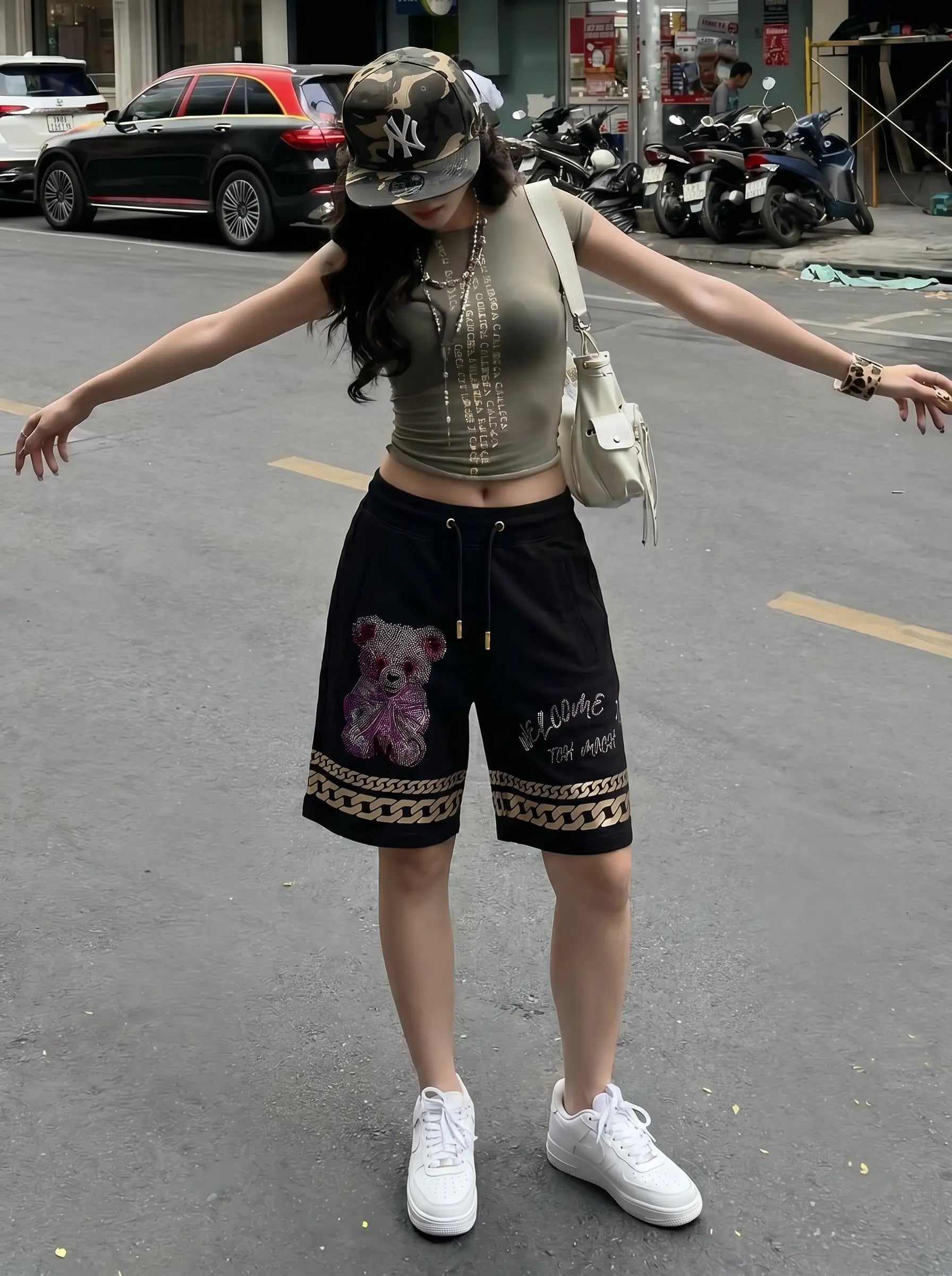 A woman stands on a city street with her arms outstretched, wearing a fitted beige crop top with vertical metallic embellishments, black drawstring shorts with a teddy bear graphic and decorative trim, a camouflage baseball cap, layered necklaces, and white sneakers. TCH Gothic Rhinestone Teddy Bear Chain Print Sweat Shorts