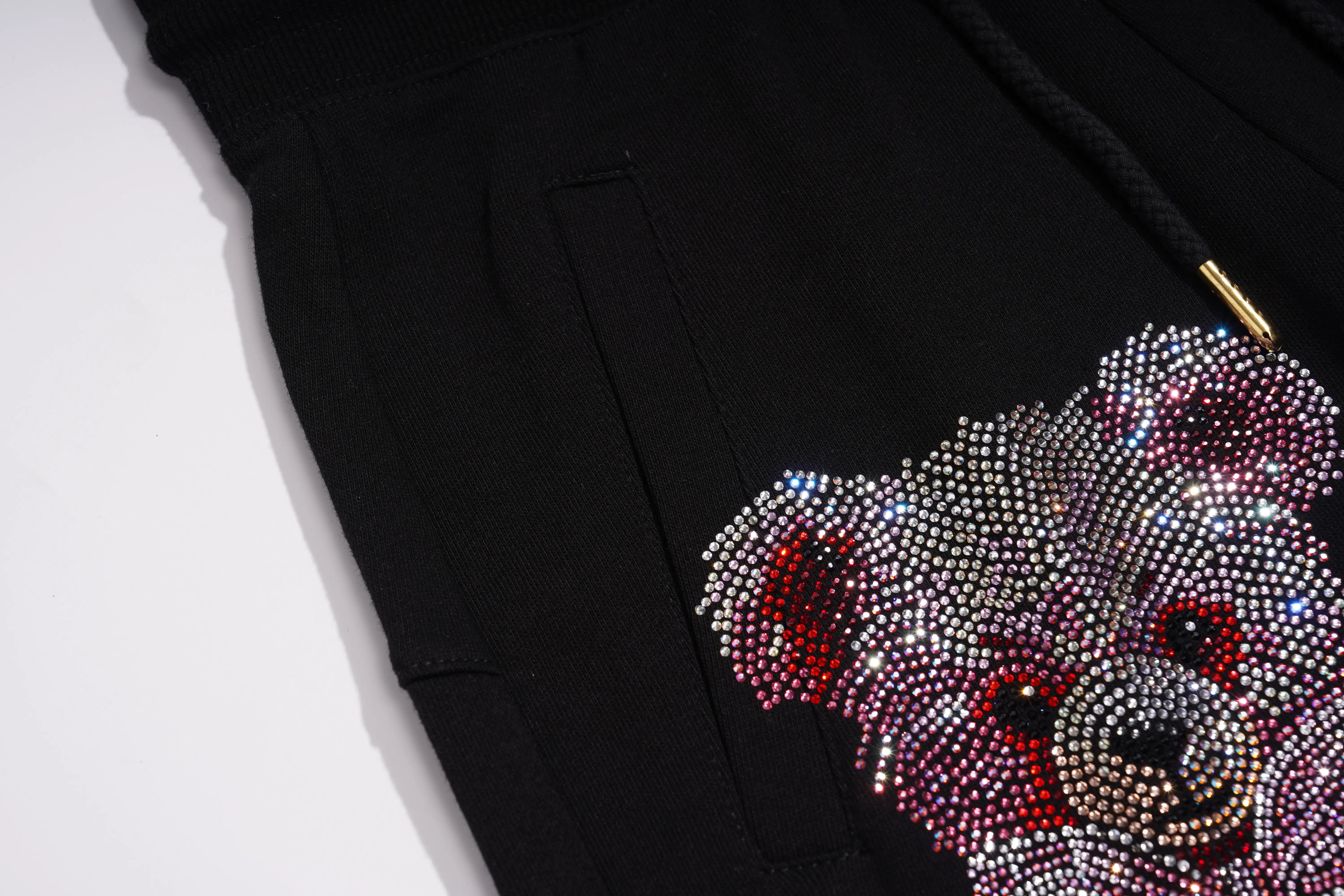Close-up of a black garment featuring a large teddy bear face embellished with sparkling rhinestones in silver, red, and pink tones, with a black drawstring and a small gold accent visible. TCH Gothic Rhinestone Teddy Bear Chain Print Sweat Shorts