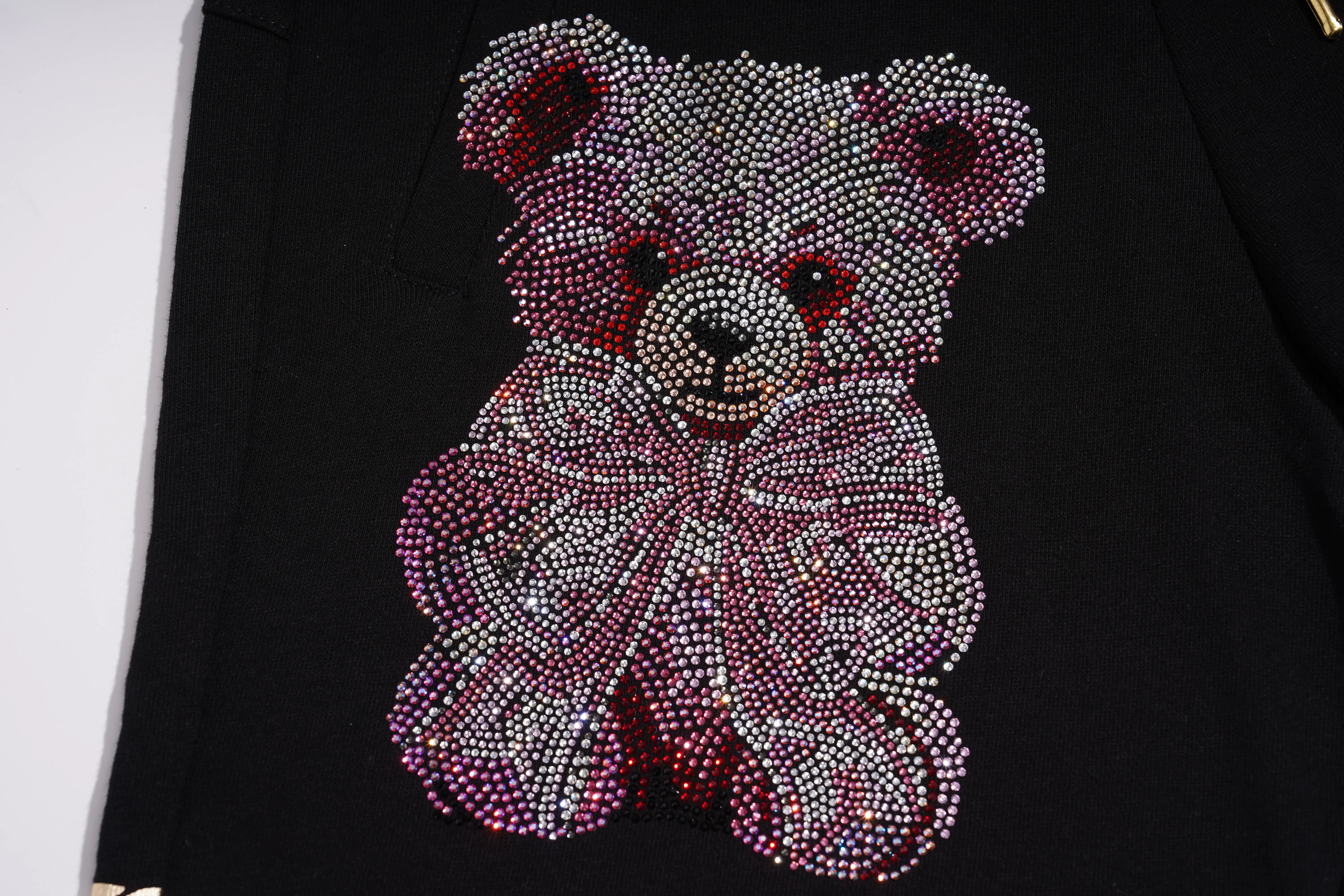 A close-up of a black garment featuring a large teddy bear design embellished with sparkling rhinestones in silver, pink, and red tones. TCH Gothic Rhinestone Teddy Bear Chain Print Sweat Shorts