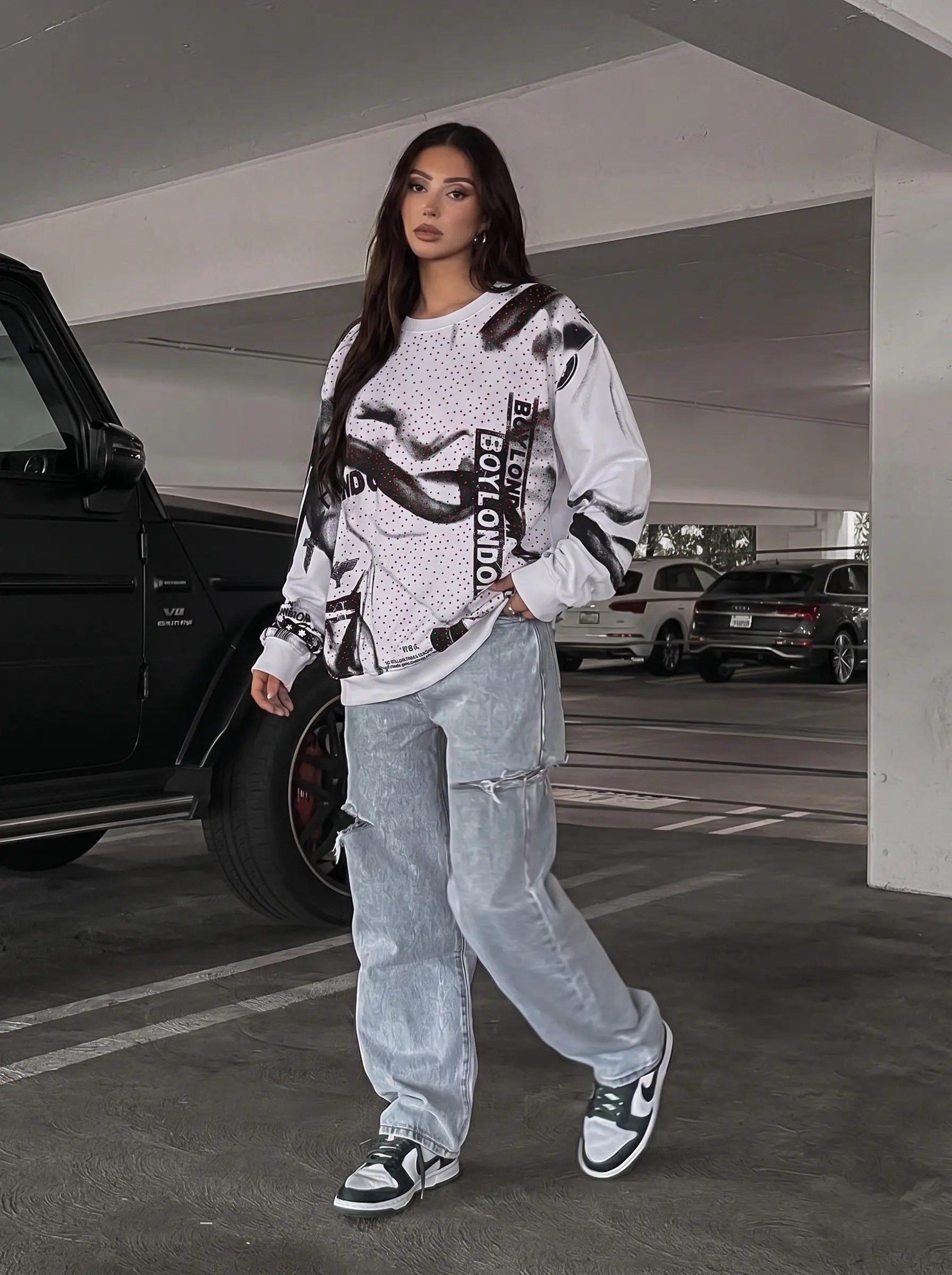 Person wearing a graphic sweatshirt and light jeans in a parking garage. TCH Graffiti Logo Print Crewneck Sweatshirt