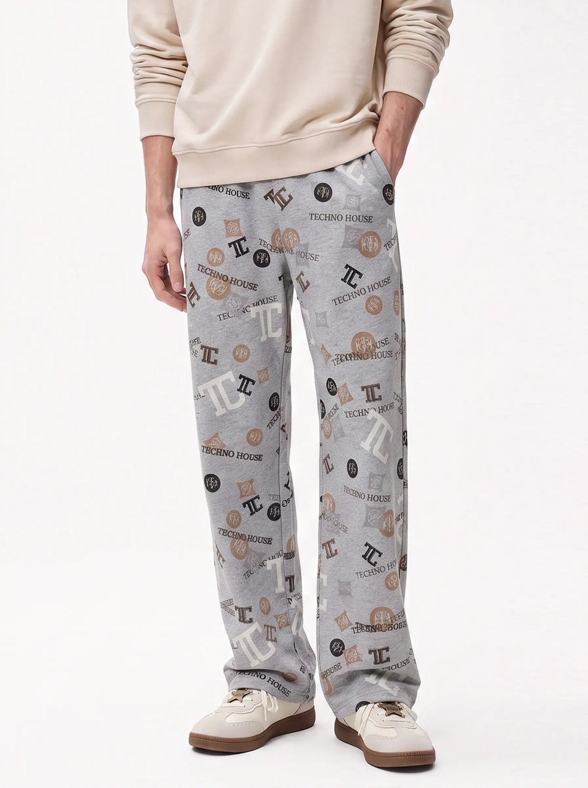 A person wearing light gray jogger-style pants with an allover printed pattern of black and brown 'TE' initials and 'TIGRENO HOUSE' text, paired with a beige sweatshirt and beige-and-brown shoes, against a plain white studio background. TCH Gray Techno Graphic Wide-Leg Sweatpants