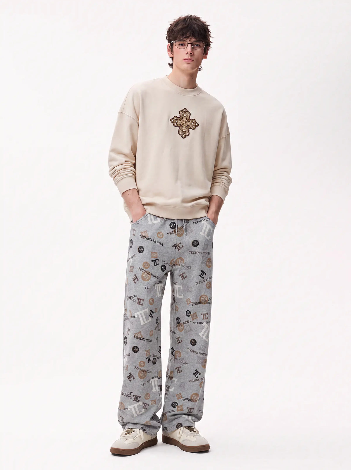 A man modeling a cream sweatshirt with a brown cross emblem on the chest and gray patterned pants, standing against a plain white studio background. TCH Gray Techno Graphic Wide-Leg Sweatpants
