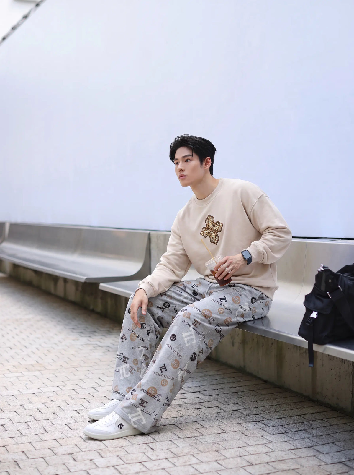 A man sitting on a concrete bench outdoors, wearing a beige crewneck sweatshirt with a gold graphic on the chest and light gray patterned sweatpants. A black backpack sits on the bench next to him. TCH Gray Techno Graphic Wide-Leg Sweatpants