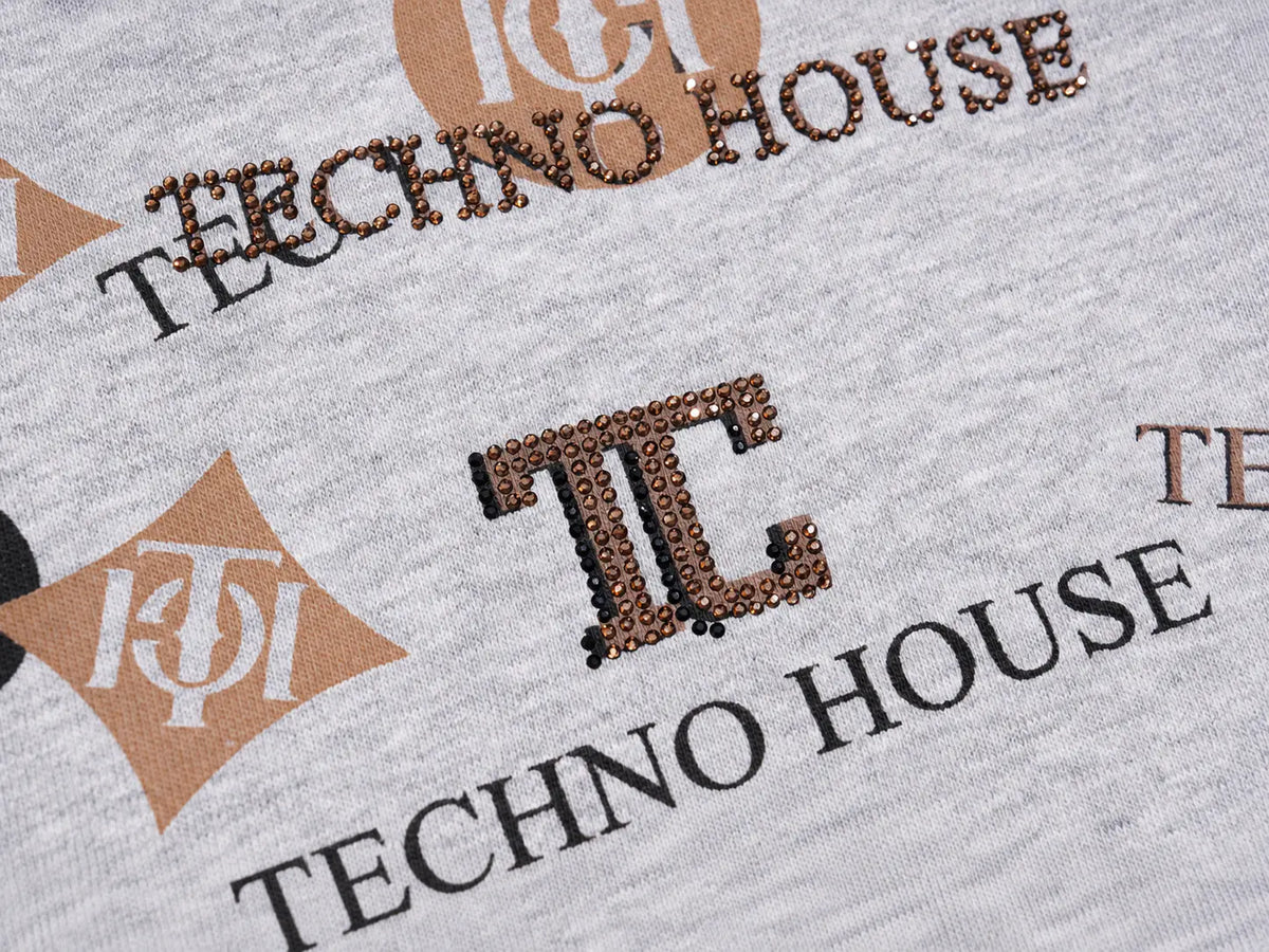 Close-up of a light heather gray garment featuring a repeating Techno House logo print in black and tan, with metallic brown sequins forming the TC monogram. TCH Gray Techno Graphic Wide-Leg Sweatpants